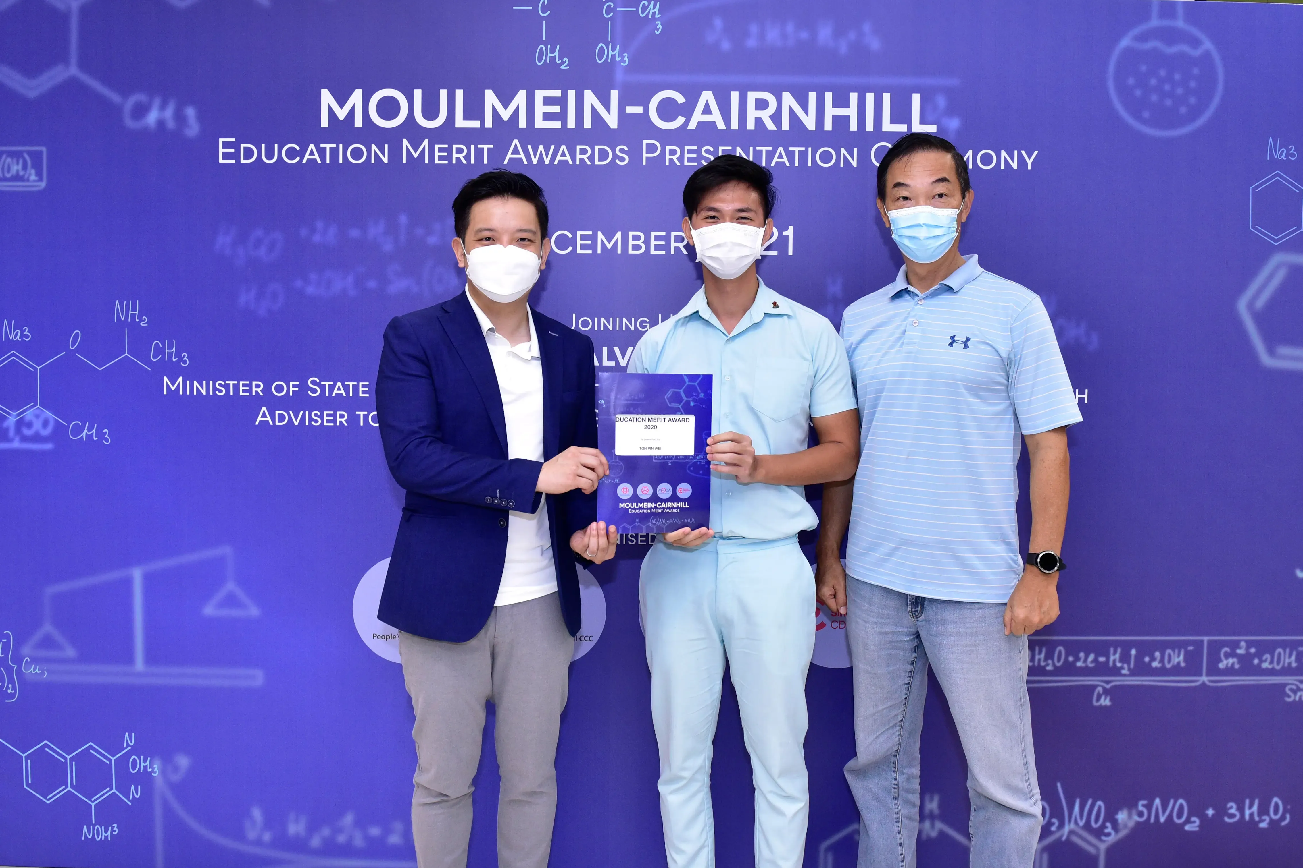 Three masked men pose at the Moulmein-Cairnhill Education Merit Awards ceremony, with the center man holding a certificate.