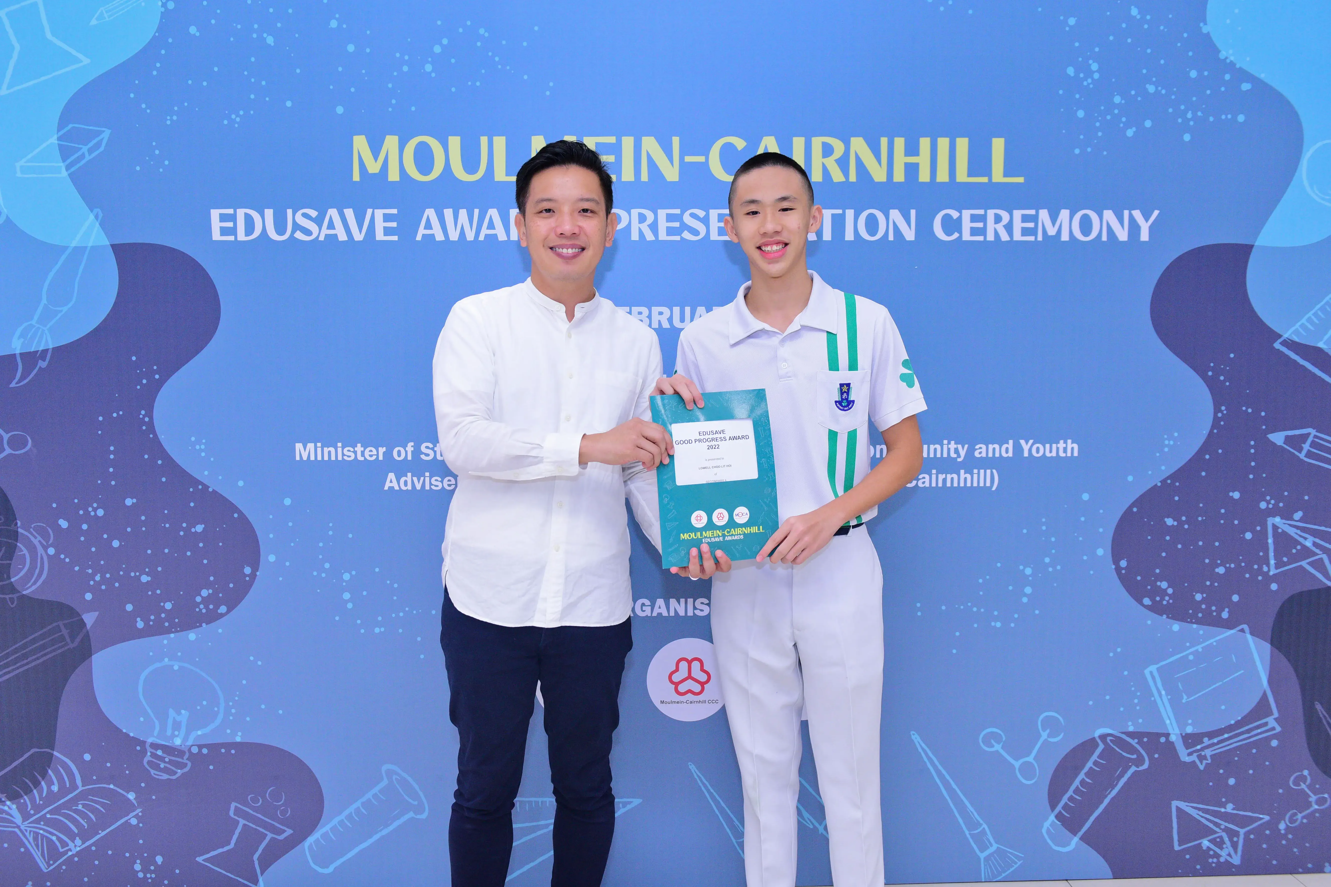 Two smiling men hold a teal Edusave Good Progress Award booklet at the Moulmein-Cairnhill award ceremony.