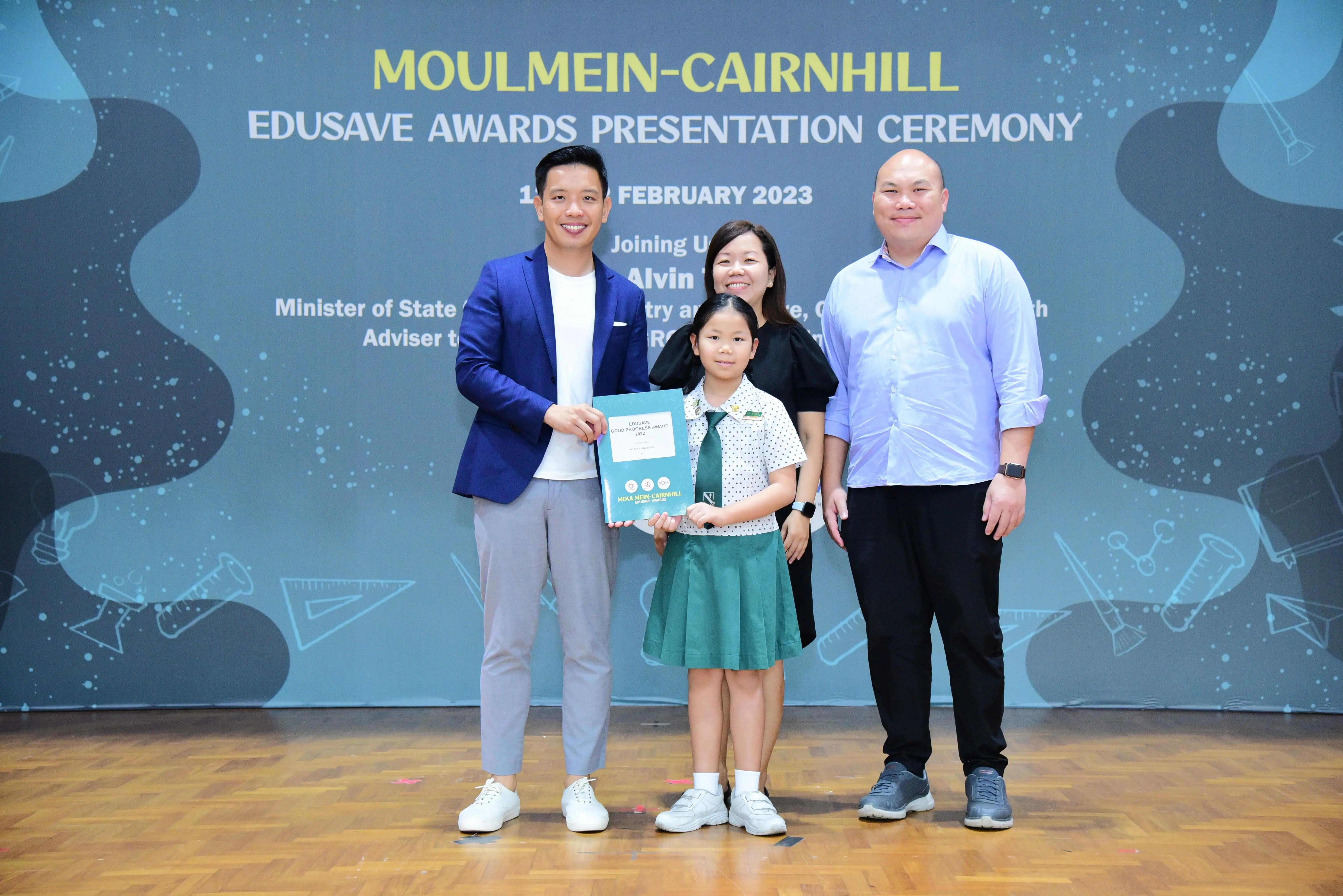 Three adults and a young girl in school uniform hold a certificate at the Moulmein-Cairnhill Edusave Awards Ceremony 2023.