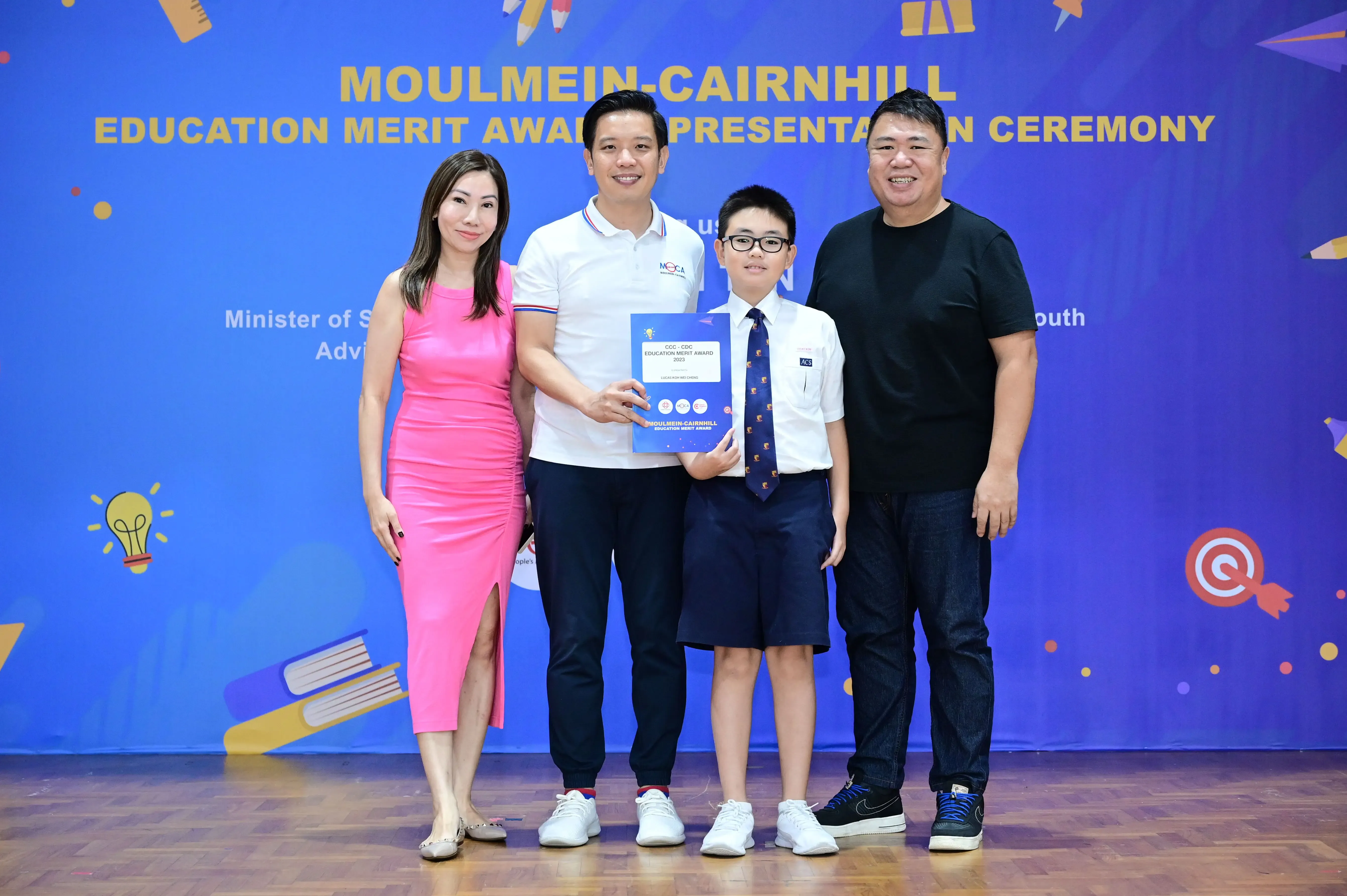 Four people pose at the Moulmein-Cairnhill Education Merit Award Presentation Ceremony, with a boy holding a certificate.