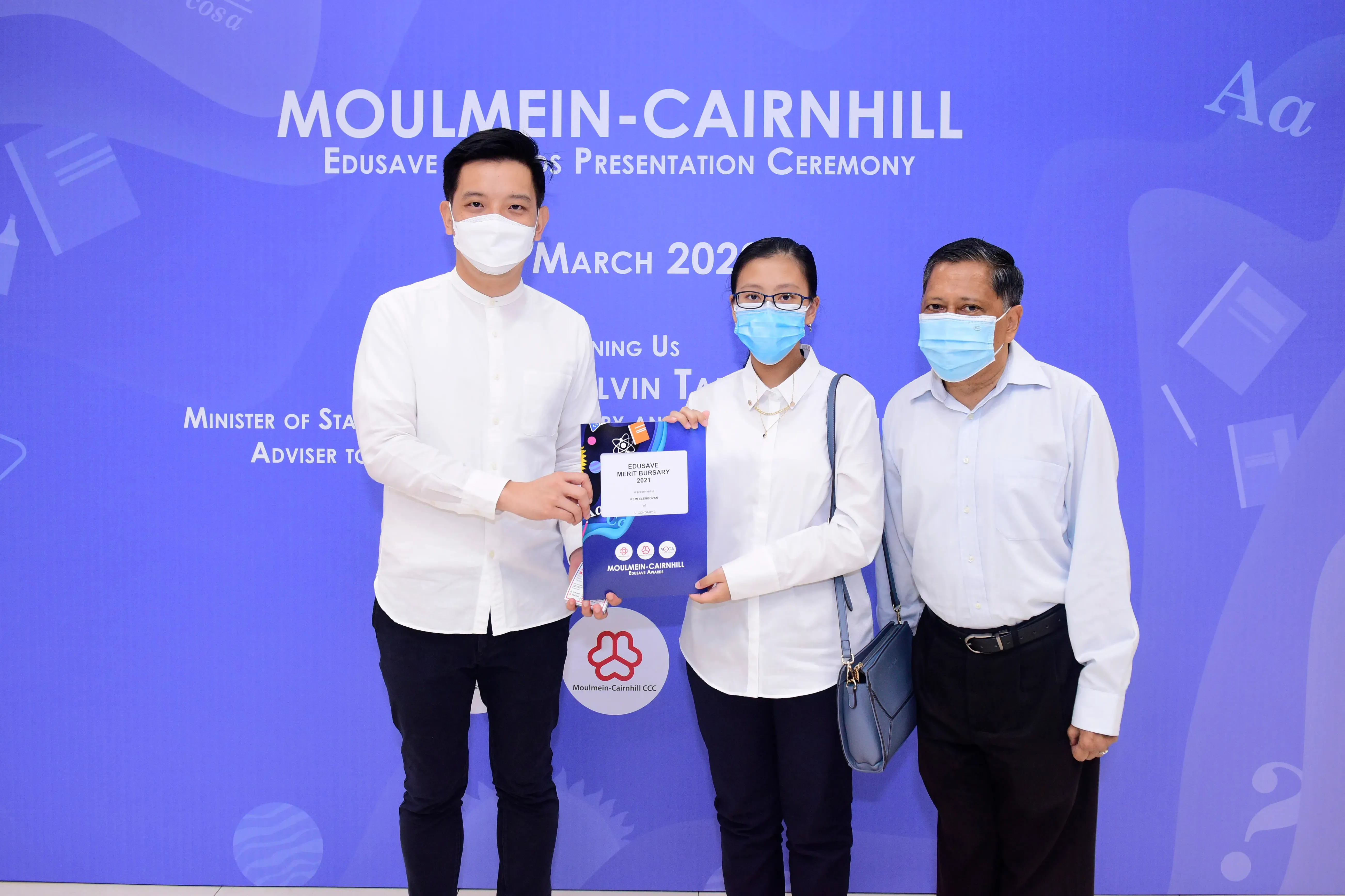 Three masked people pose at the Moulmein-Cairnhill Edusave Awards Presentation Ceremony, holding a Merit Bursary folder.