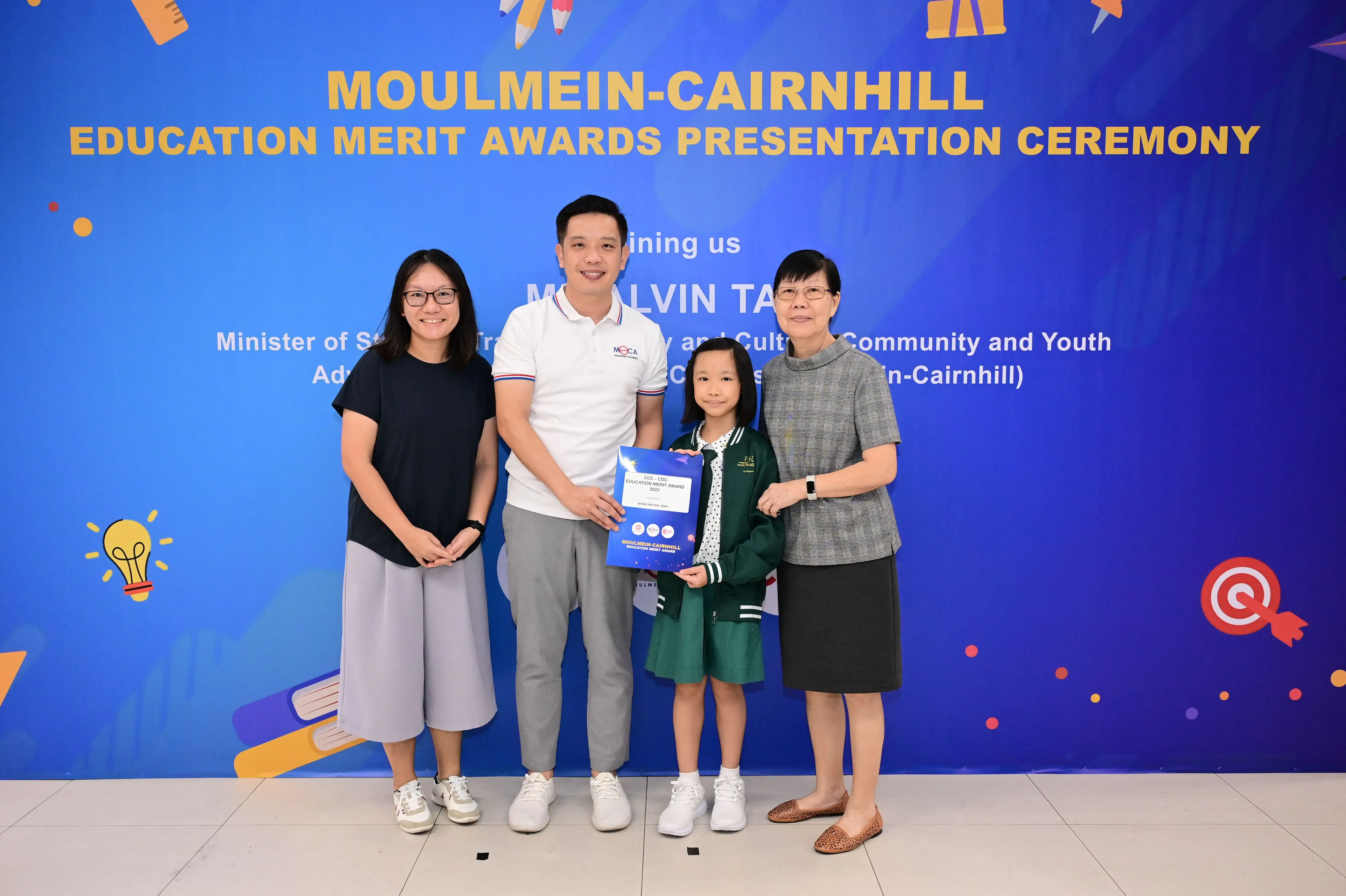 Three adults and a young girl hold an award certificate at the Moulmein-Cairnhill Education Merit Awards Presentation Ceremony.