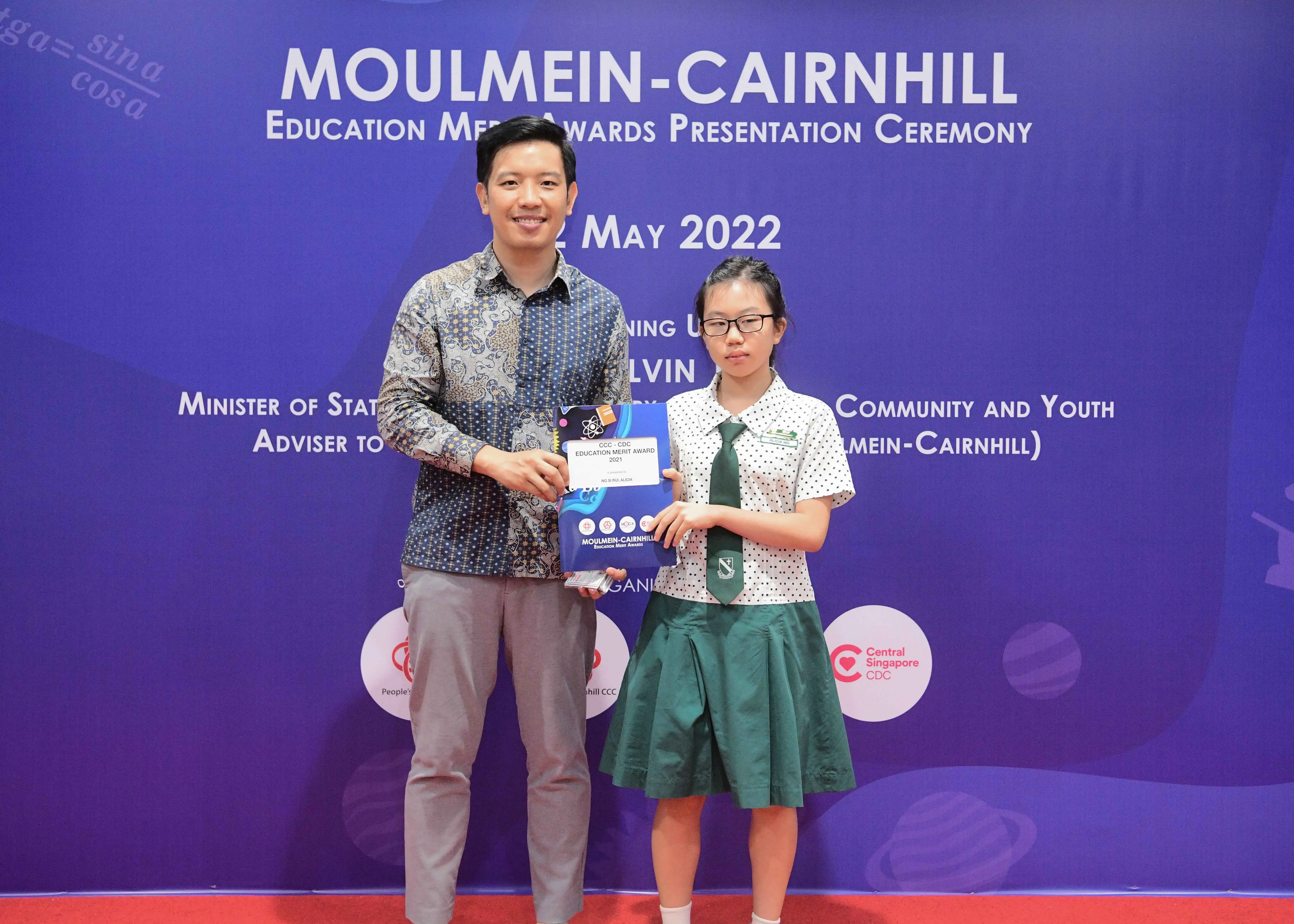 A man and a young girl in school uniform hold an award at the Moulmein-Cairnhill Education Merit Awards ceremony, May 2022.