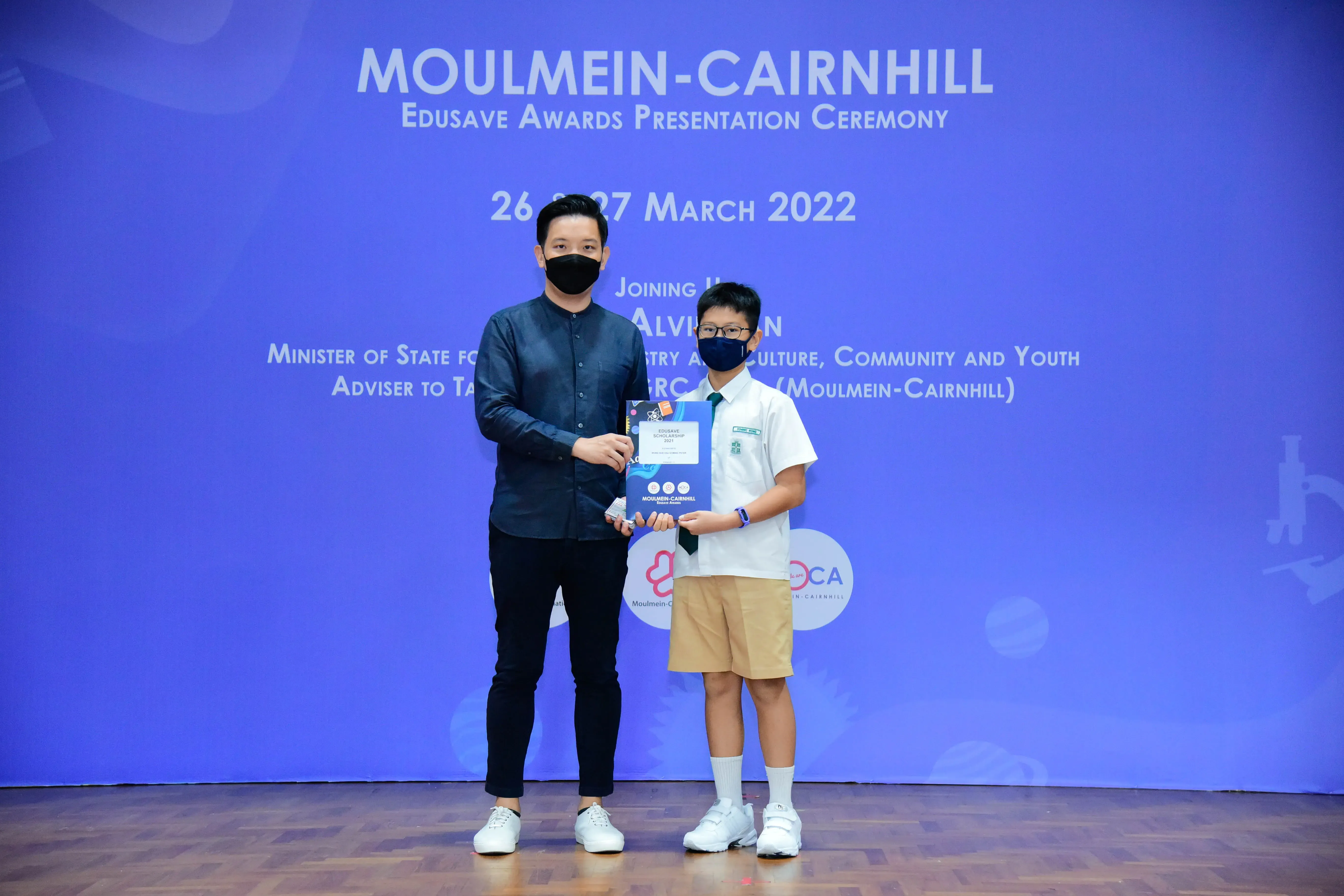An adult and a uniformed student hold an award certificate at the Moulmein-Cairnhill Edusave Awards Ceremony, March 2022.