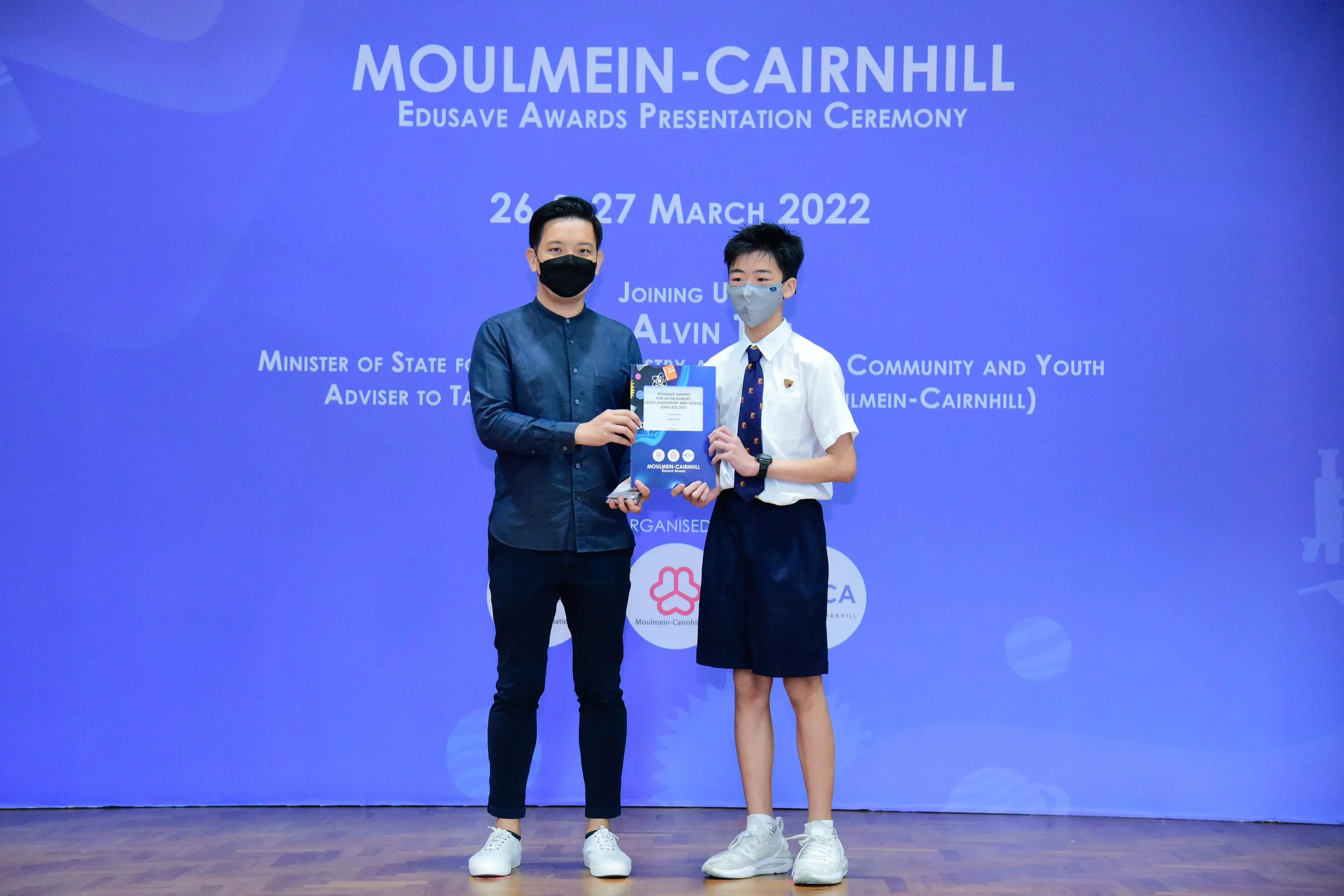 Two masked individuals hold an award certificate on stage at the Moulmein-Cairnhill Edusave Awards Ceremony, March 2022.