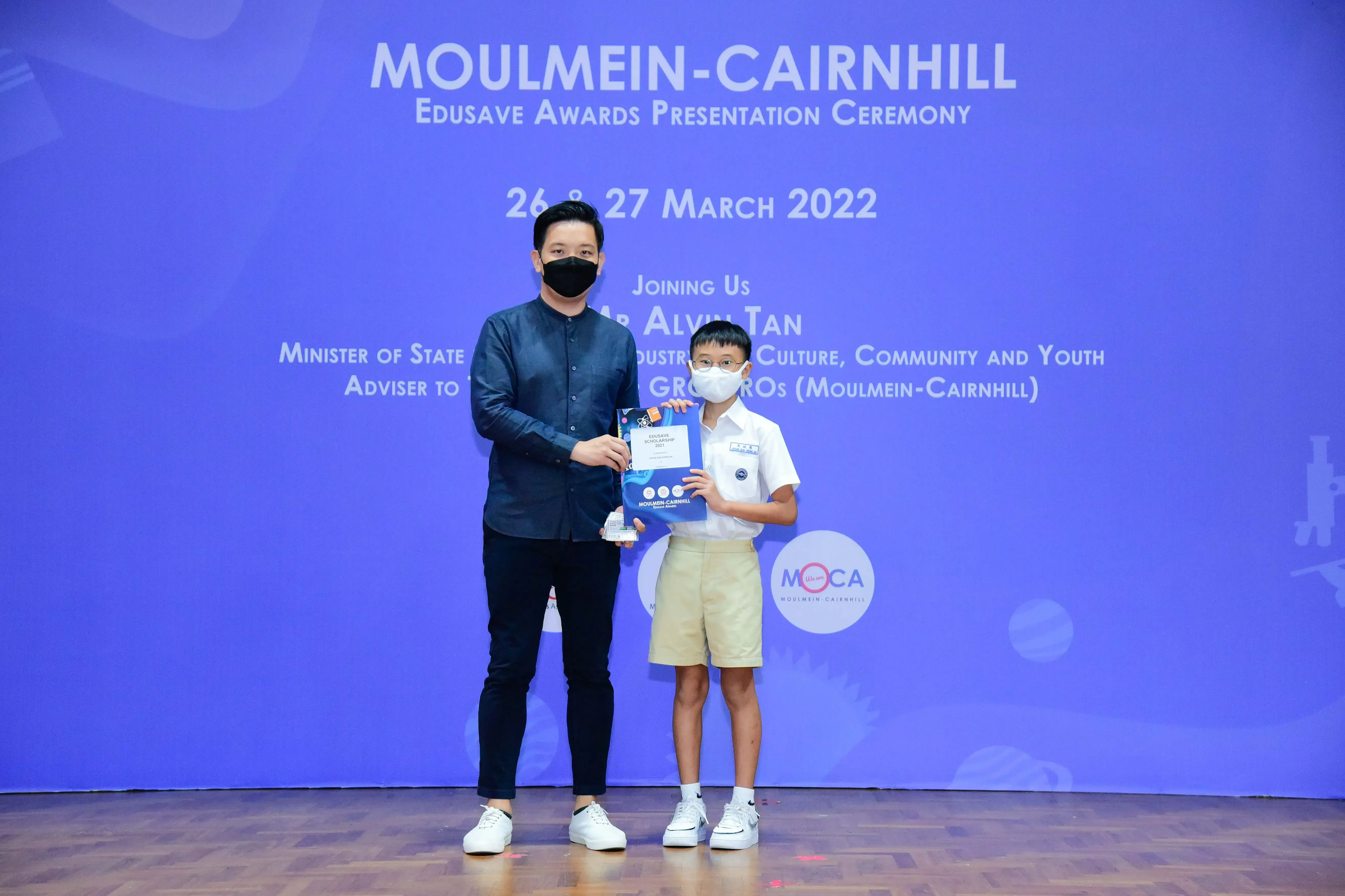An adult and a young student in school uniform hold an award at the Moulmein-Cairnhill Edusave Awards Ceremony, March 2022.