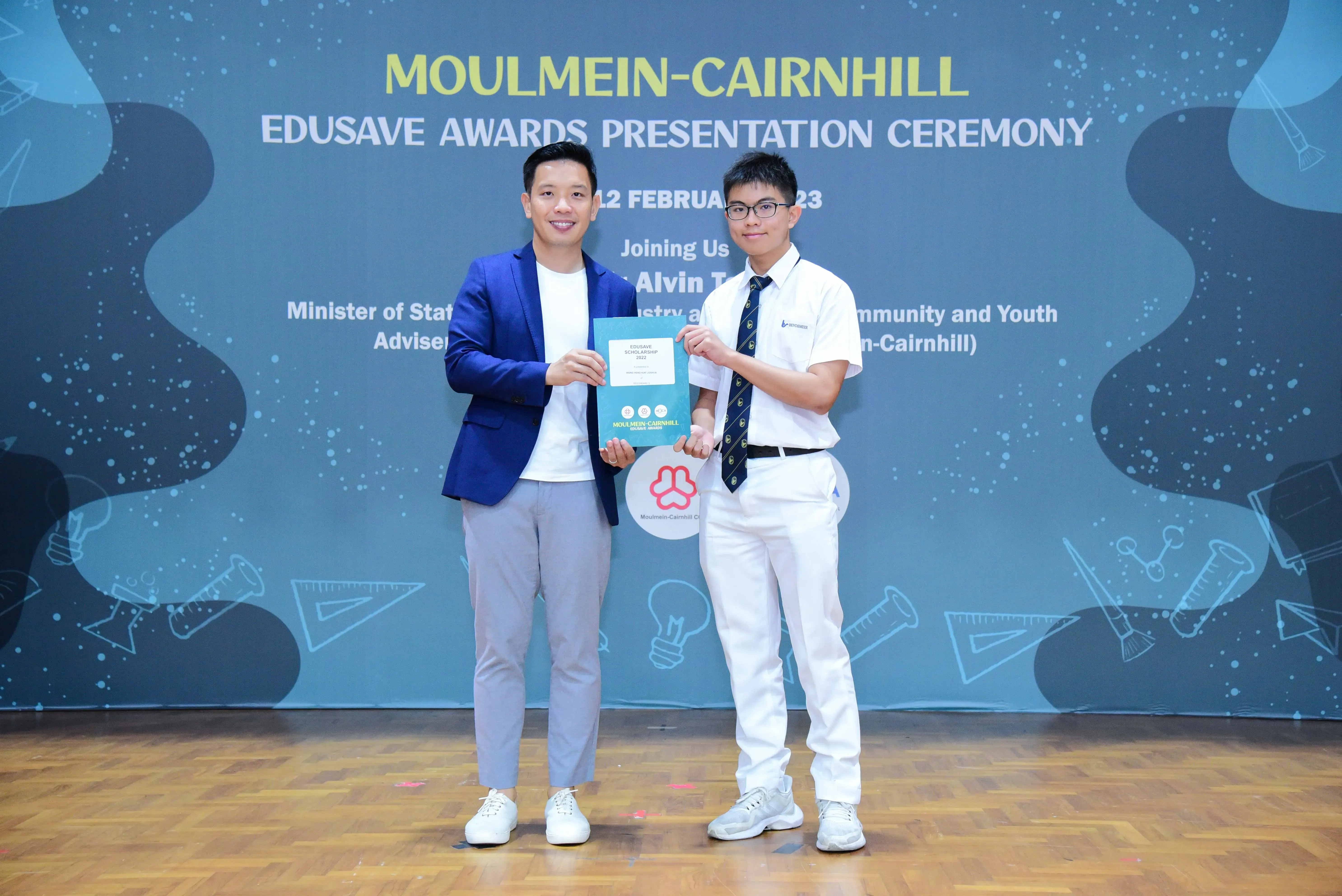 Two men hold an award certificate on stage at the Moulmein-Cairnhill Edusave Awards Presentation Ceremony.