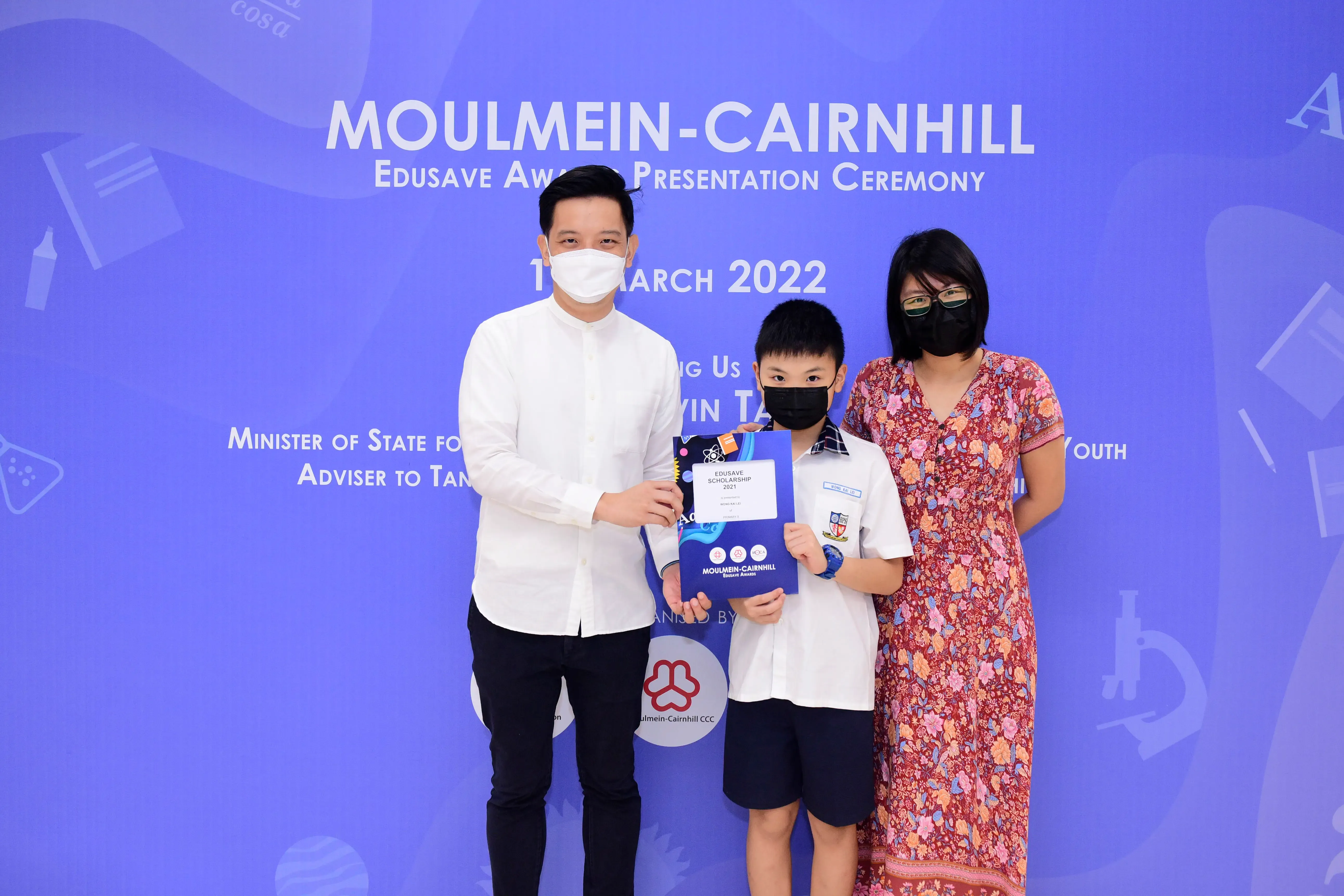 Three masked people pose at Moulmein-Cairnhill Edusave Award Presentation Ceremony in March 2022, boy holds certificate.