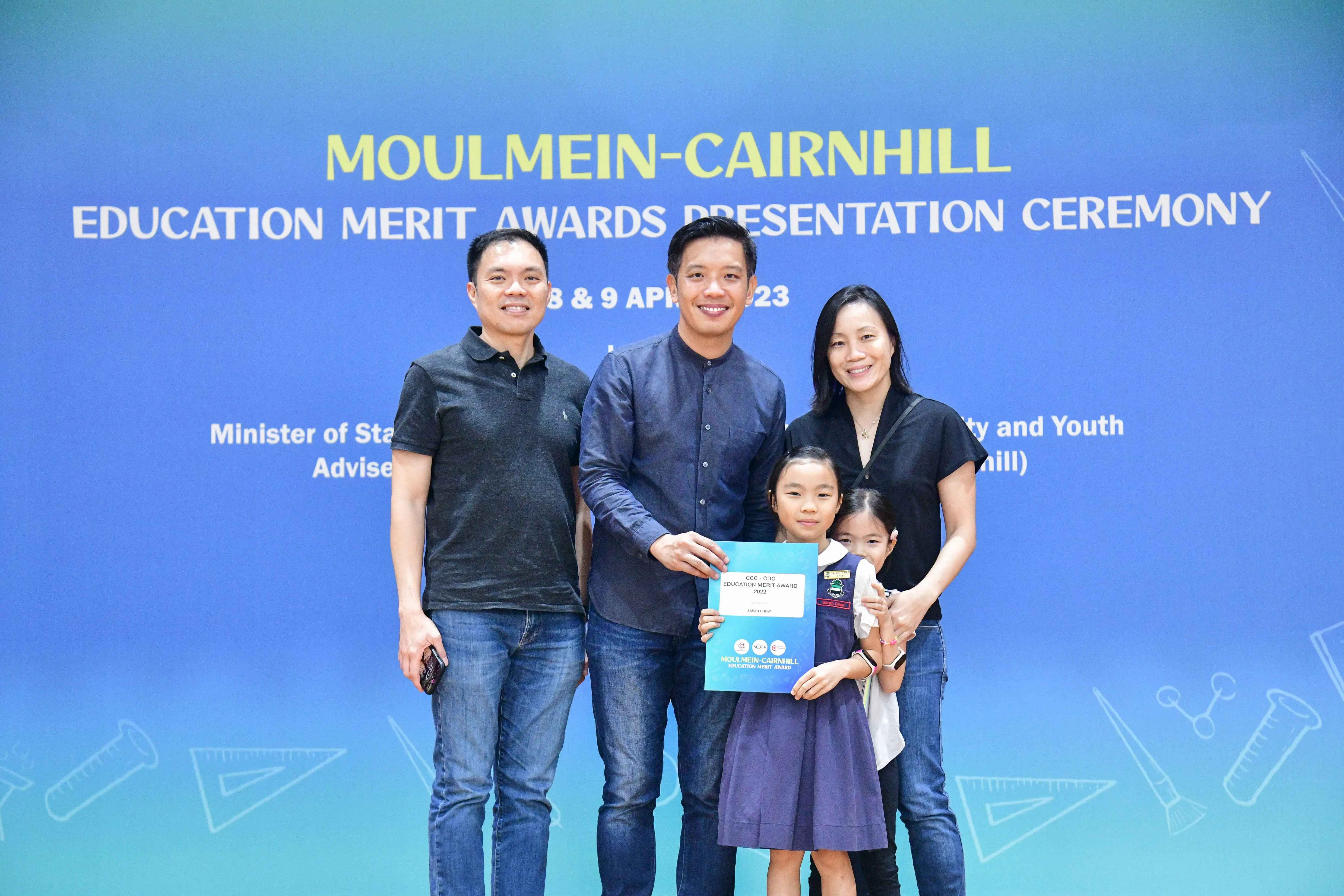 Three adults and two young girls pose holding an award certificate at the Moulmein-Cairnhill Education Merit Awards ceremony.