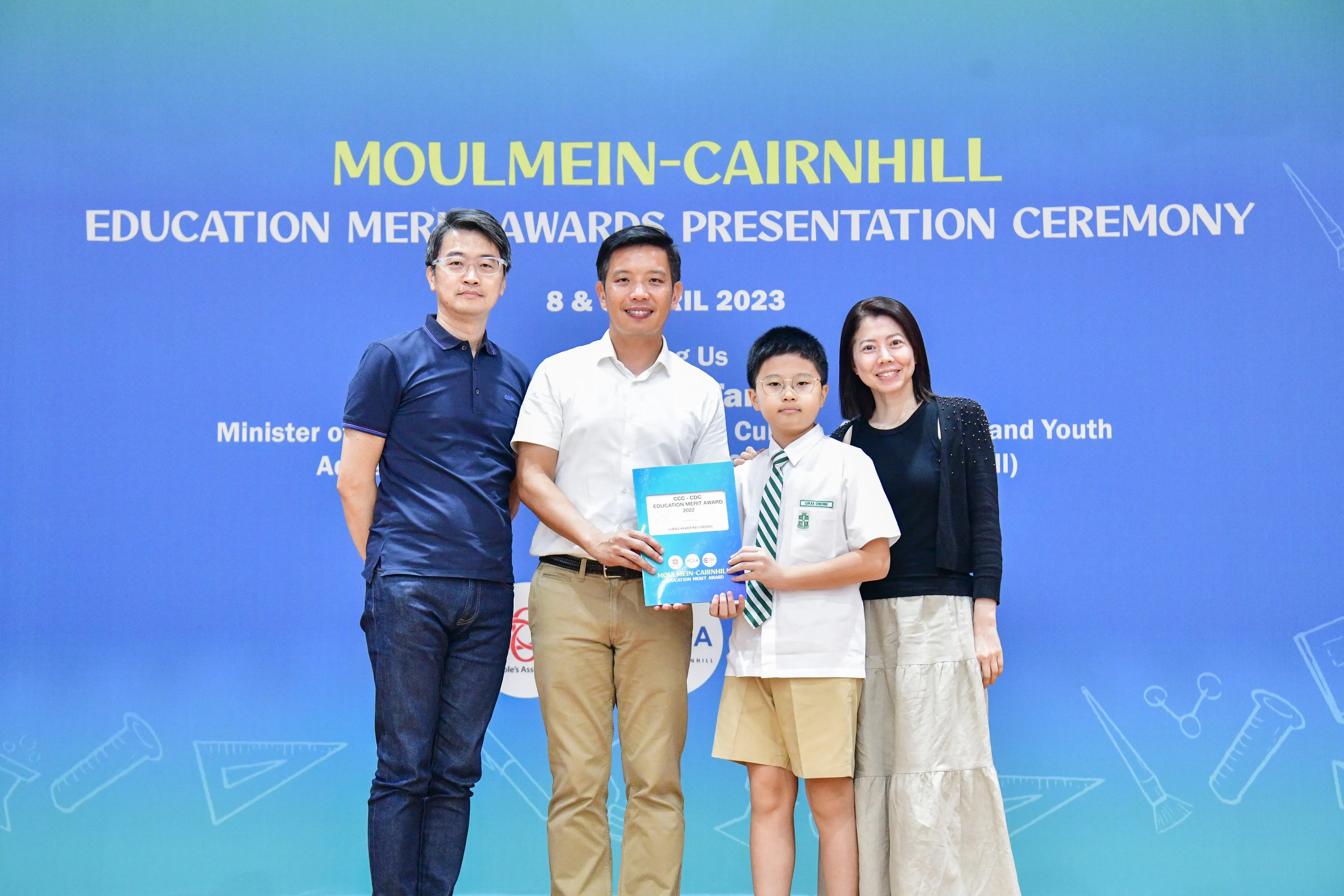 Four people pose before a Moulmein-Cairnhill Education Merit Awards Presentation Ceremony banner, a boy holding an award.