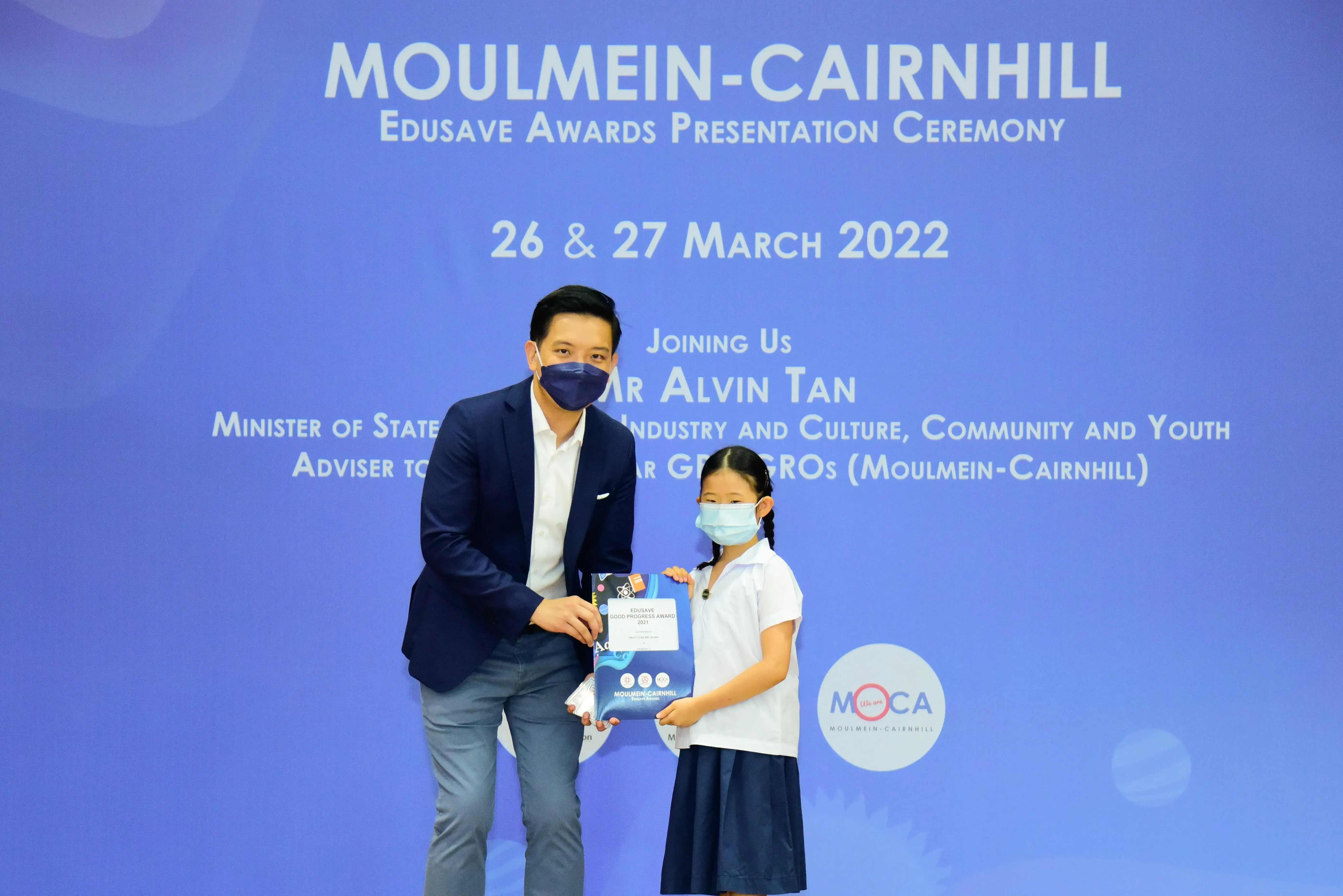 A masked man in a blazer presents an award to a masked young girl at the Moulmein-Cairnhill Edusave Awards Ceremony 2022.