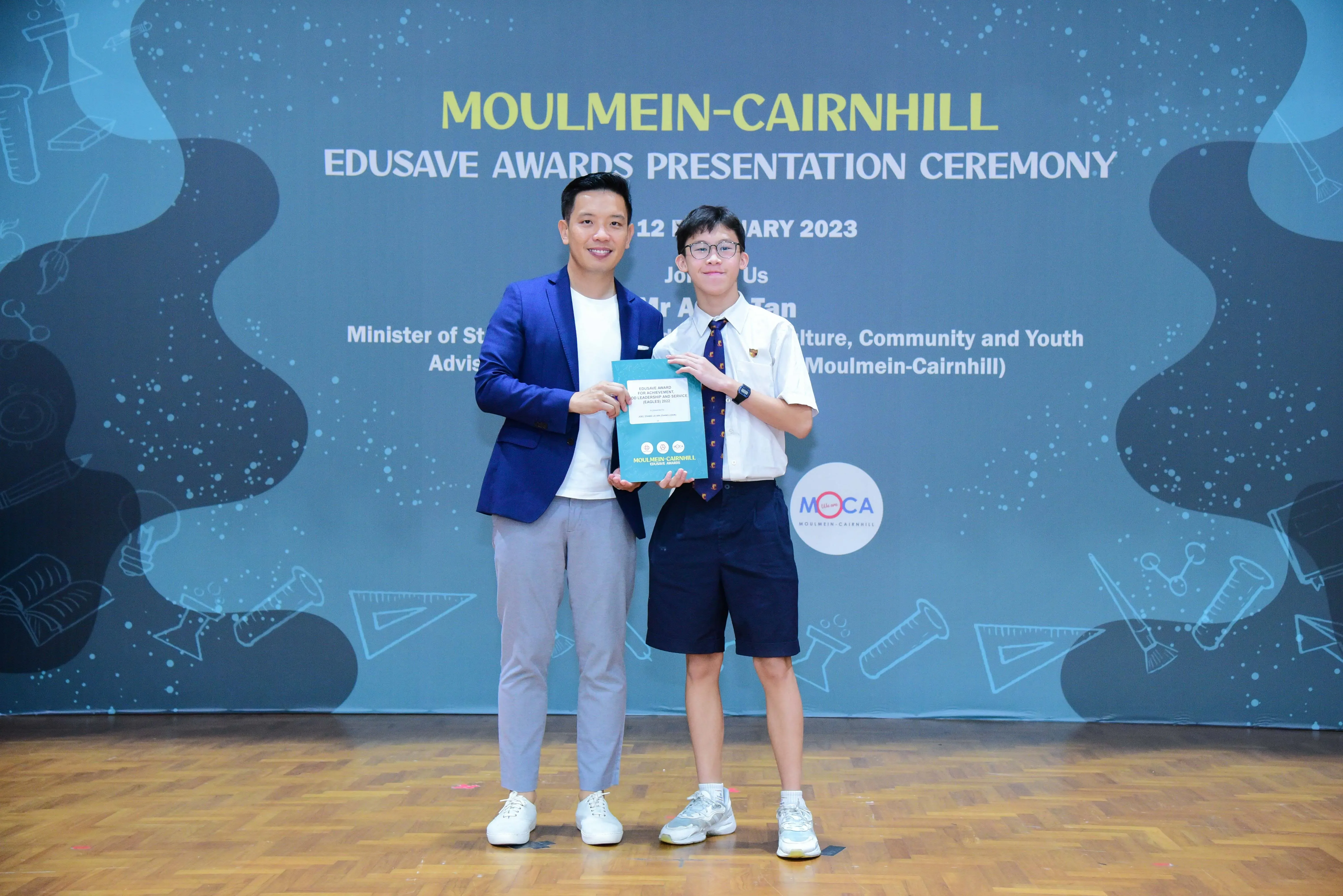 A man in a blue blazer and a student in uniform hold a certificate at the Moulmein-Cairnhill Edusave Awards Ceremony 2023.
