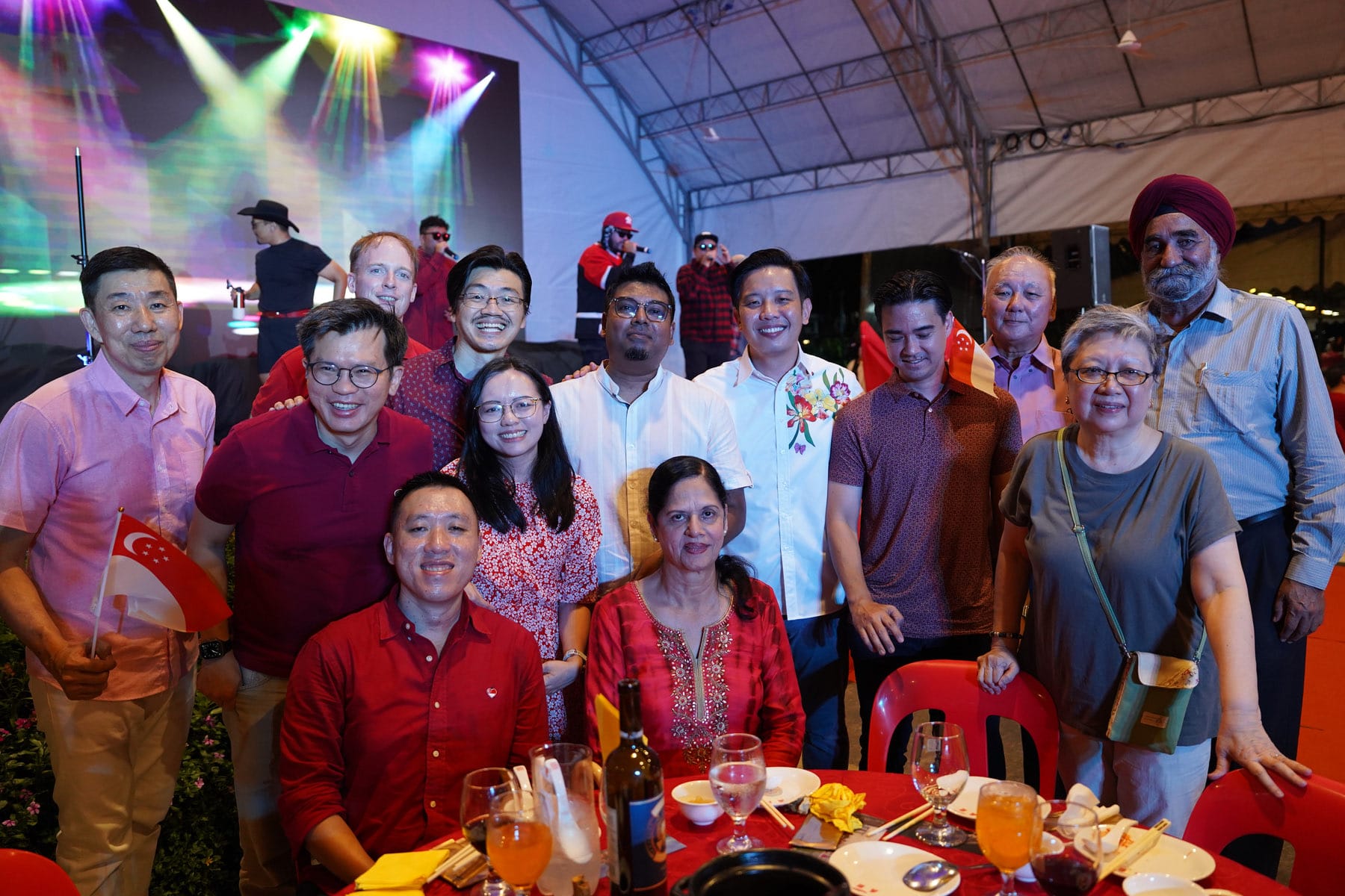 A large group of smiling people pose together at an outdoor dinner event with colorful stage lights in the background.