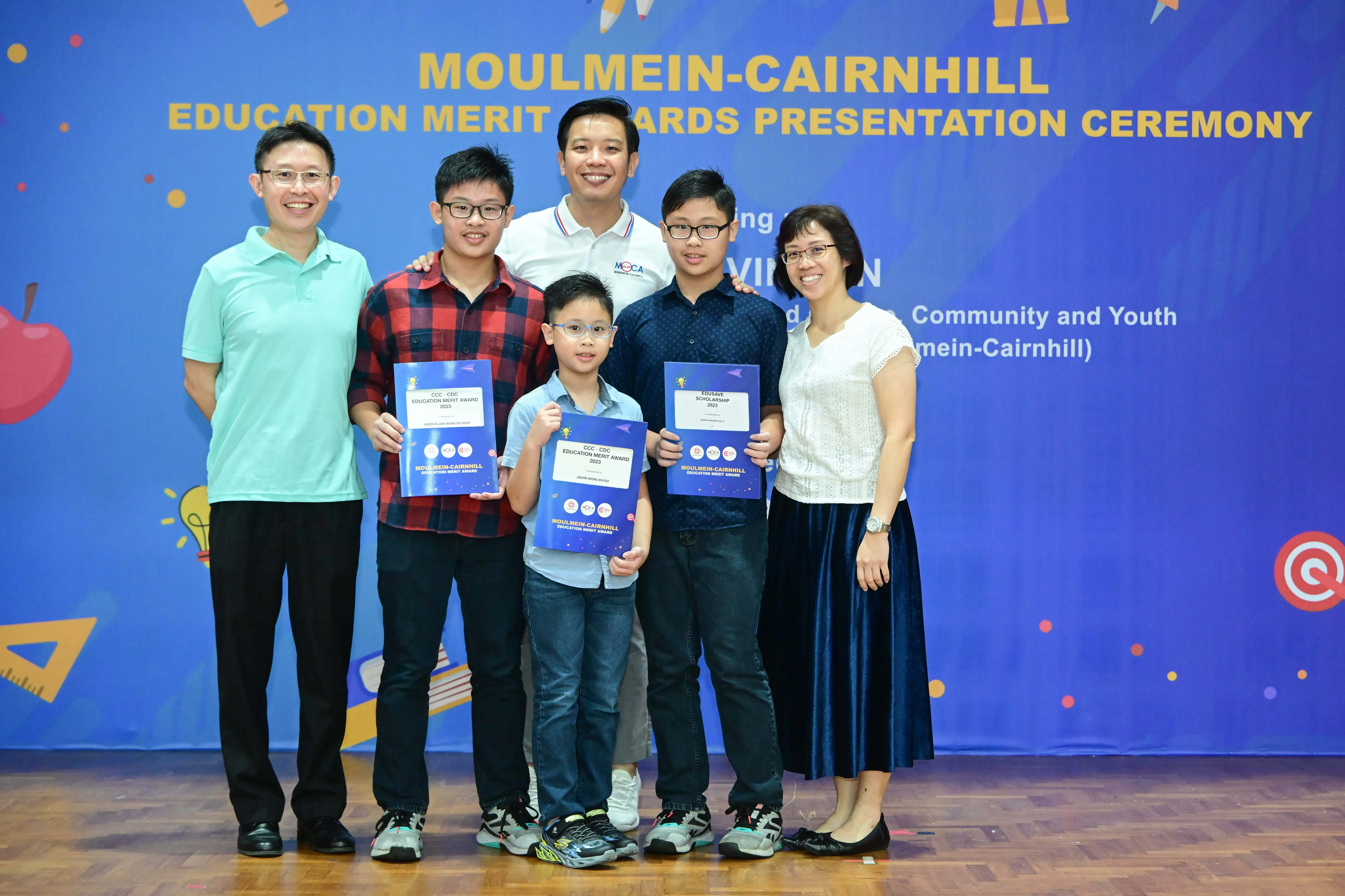 Six people pose in front of a blue Moulmein-Cairnhill Education Merit Awards banner, with three boys holding award certificates.