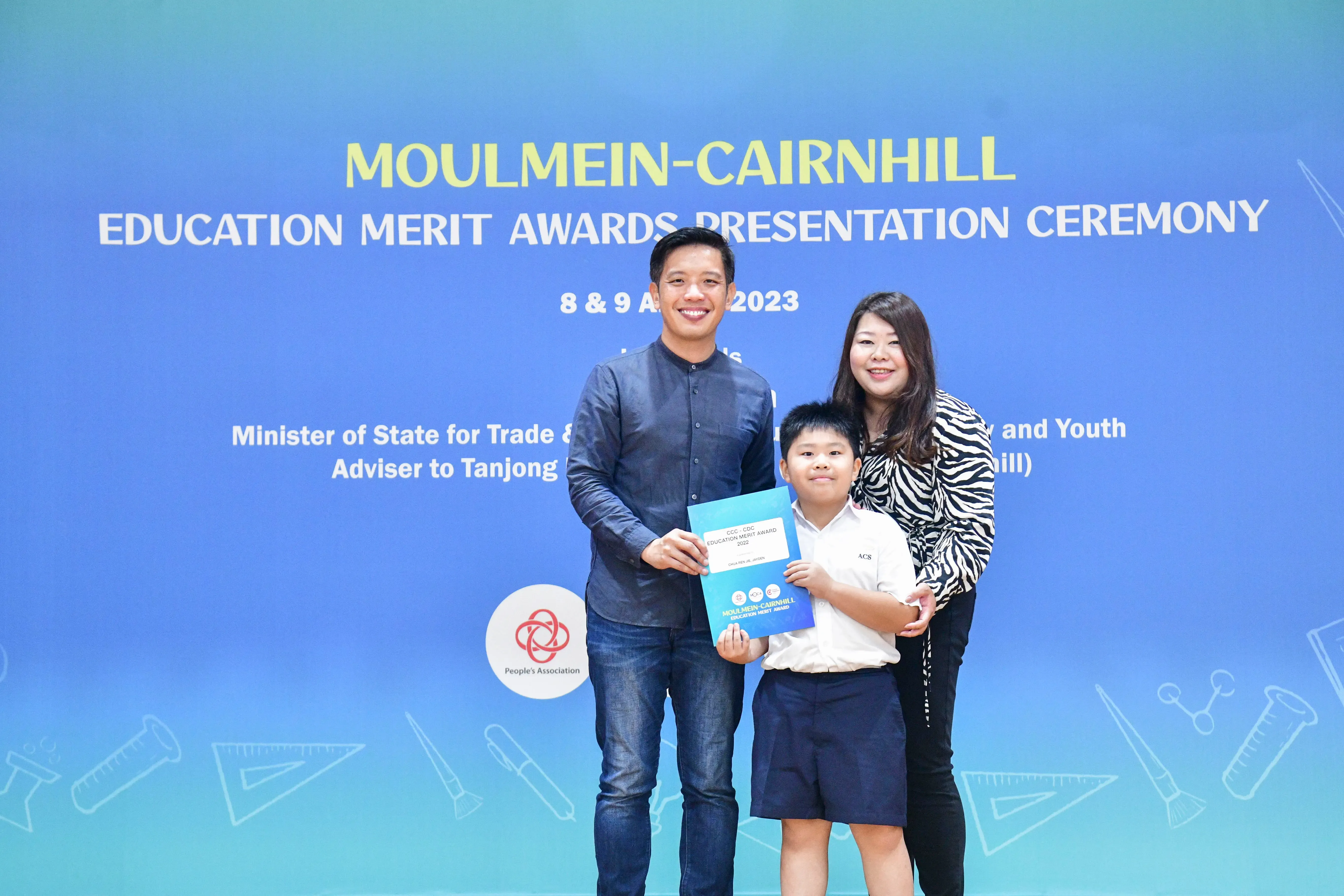 Two adults and a boy holding an award certificate pose in front of a Moulmein-Cairnhill Education Merit Awards banner.