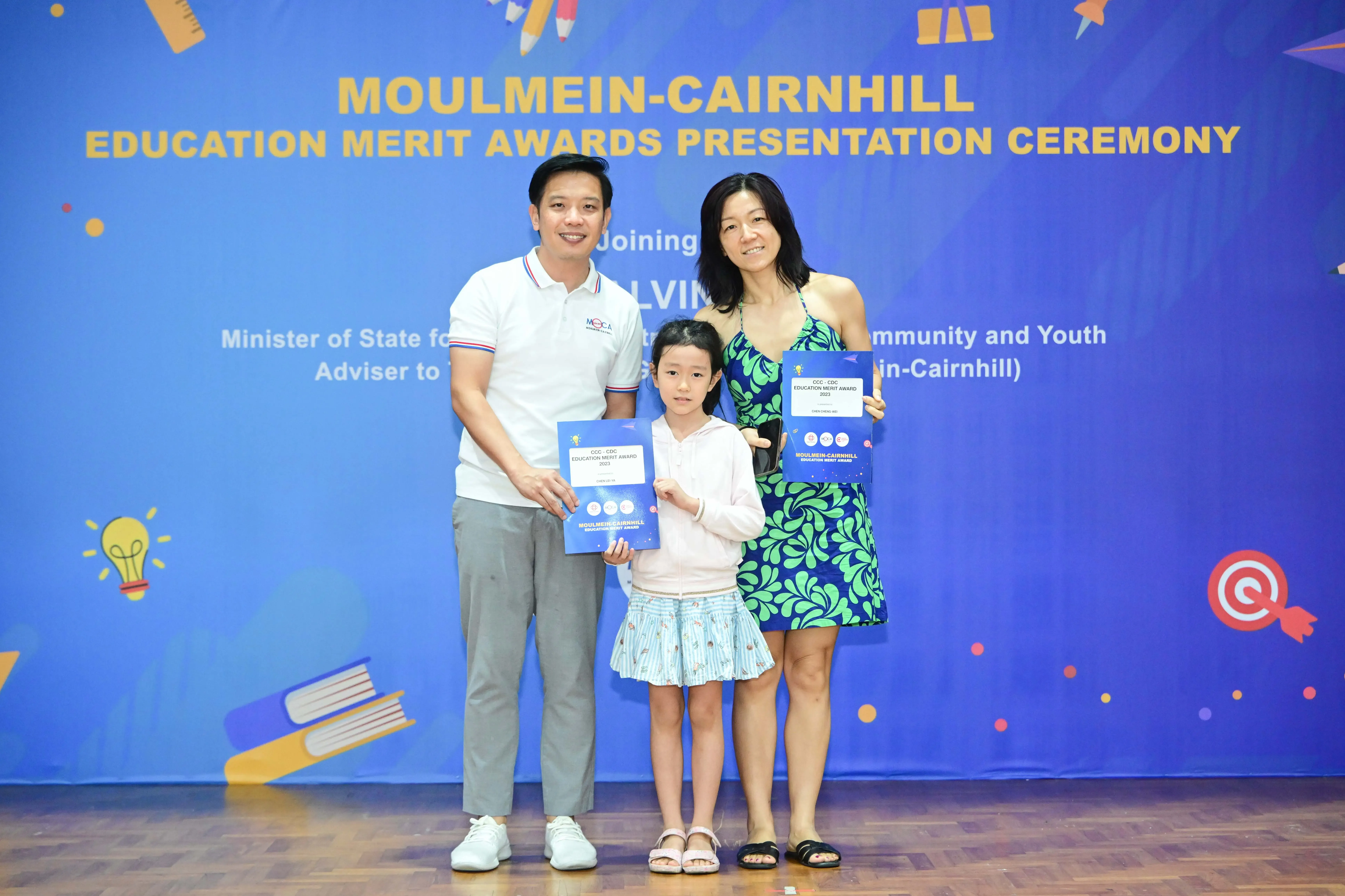 Two adults and a young girl hold award certificates in front of a Moulmein-Cairnhill Education Merit Awards banner.