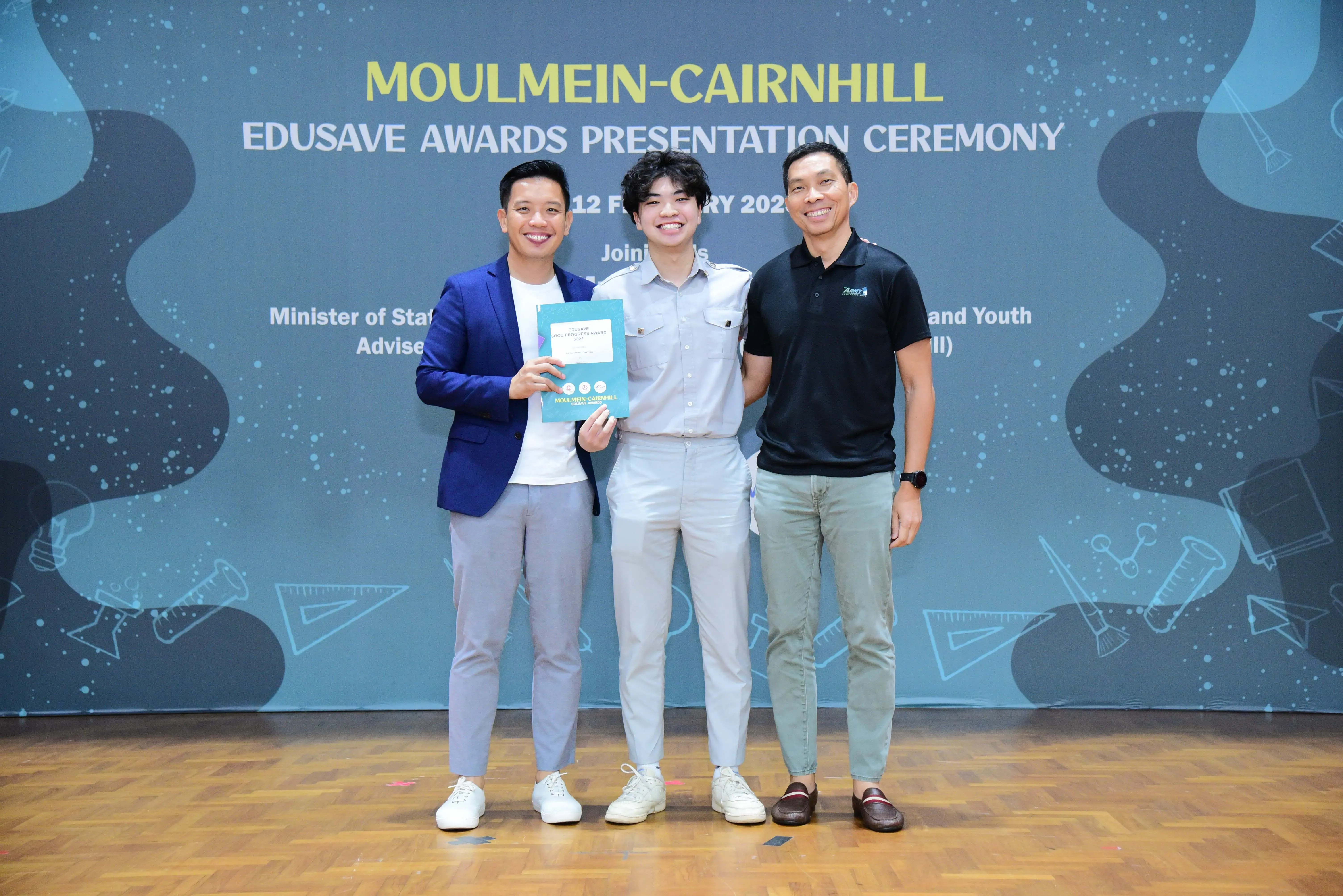 Three smiling men pose at the Moulmein-Cairnhill Edusave Awards Presentation Ceremony, the center person holding a certificate.