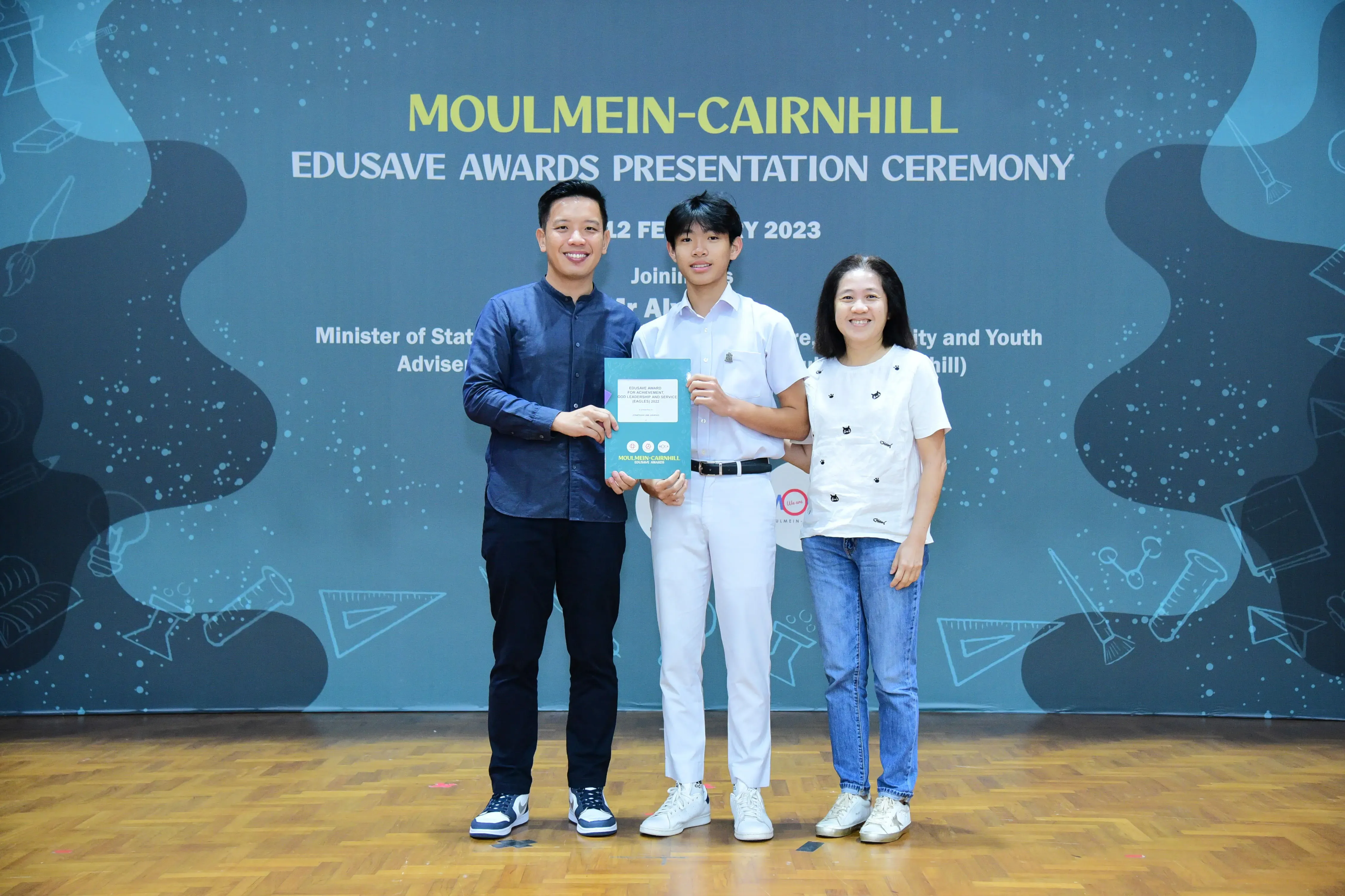 Three people smile on stage at the Moulmein-Cairnhill Edusave Awards Presentation Ceremony, holding a teal certificate.