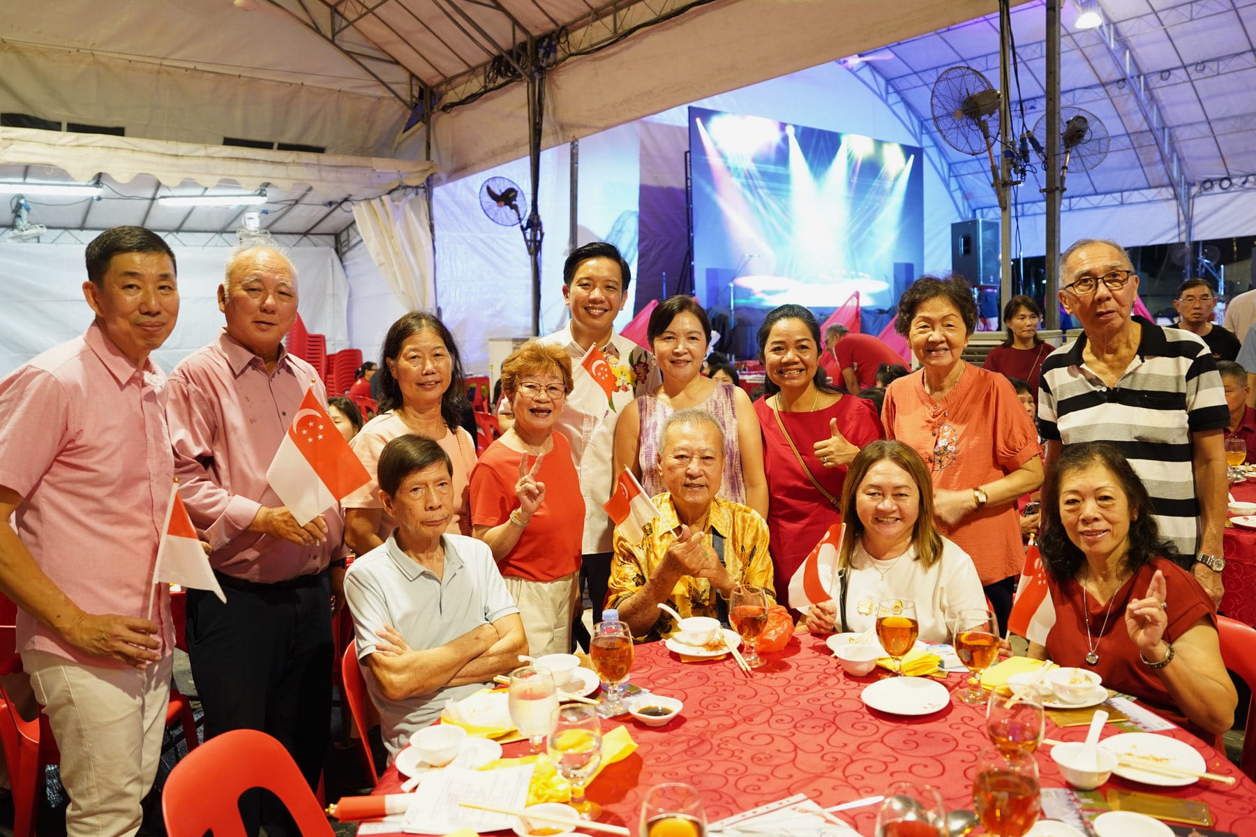 Group of people in red outfits holding Singapore flags, posing at a festive outdoor dinner event under a tent.