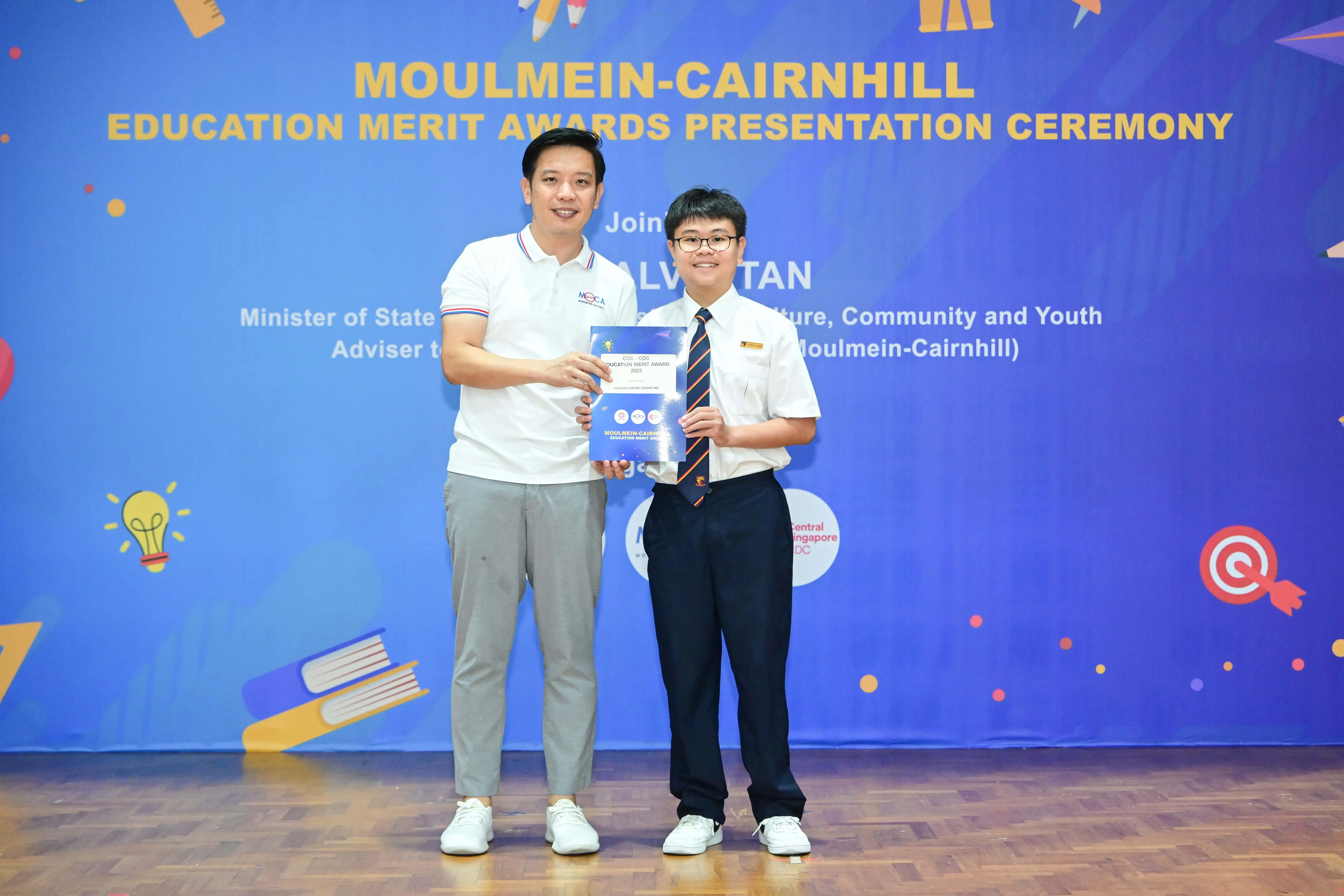 Two people hold an award certificate in front of a Moulmein-Cairnhill Education Merit Awards Presentation Ceremony banner.