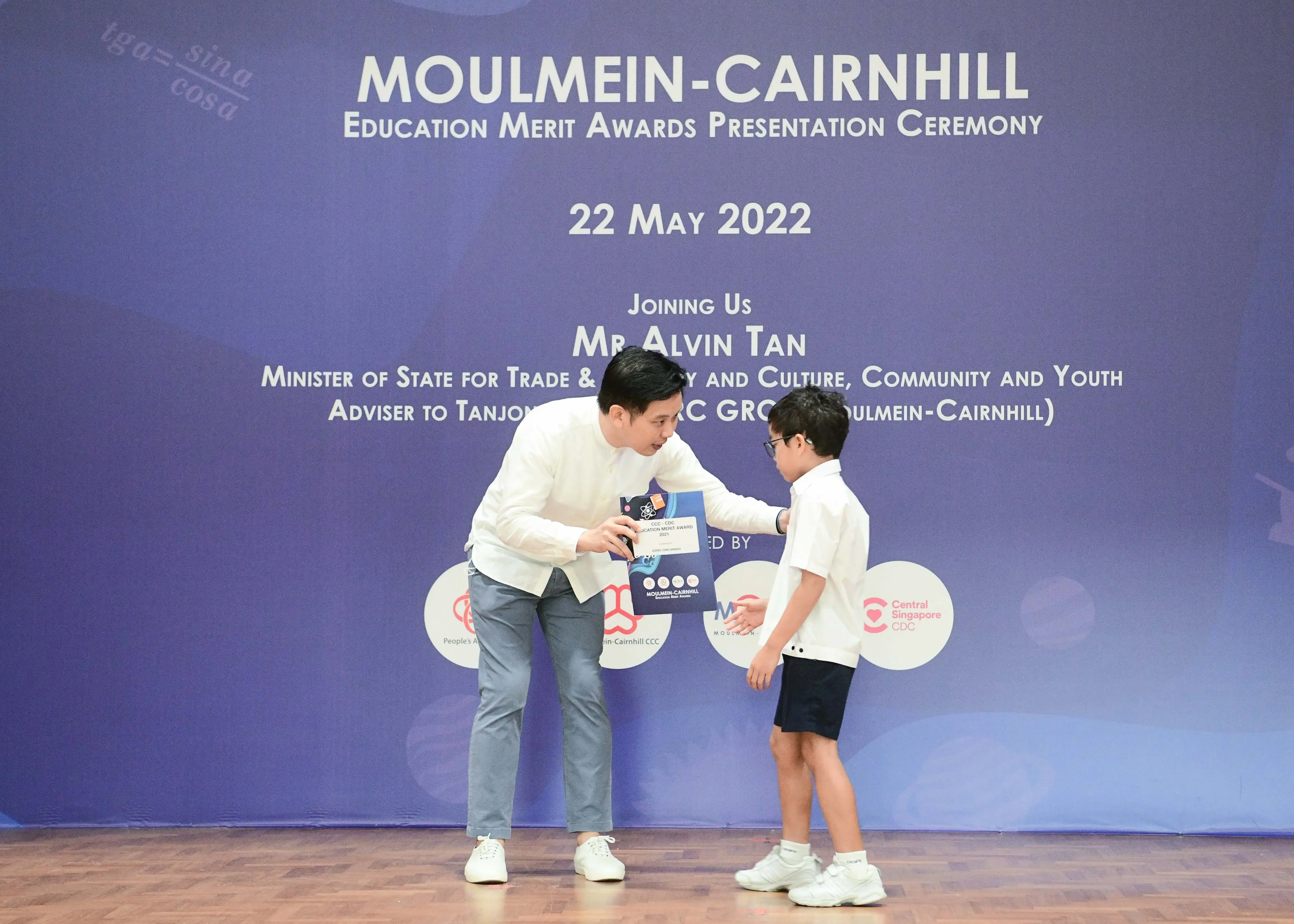 An adult in white hands an award to a young boy on stage at the Moulmein-Cairnhill Education Merit Awards, May 2022.