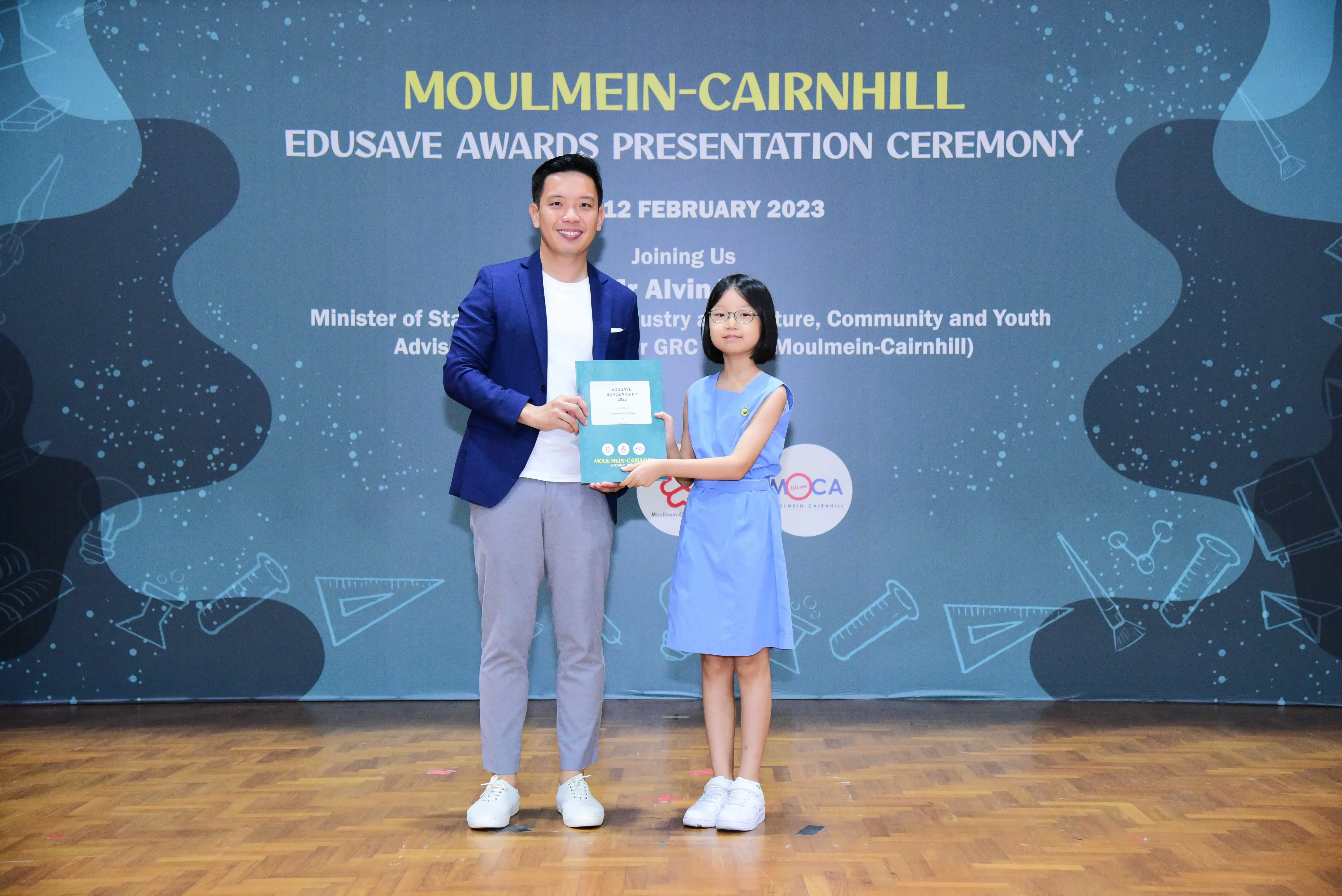 A man in a blue blazer and a young girl in a blue dress hold a certificate at the Moulmein-Cairnhill Edusave Awards Ceremony.