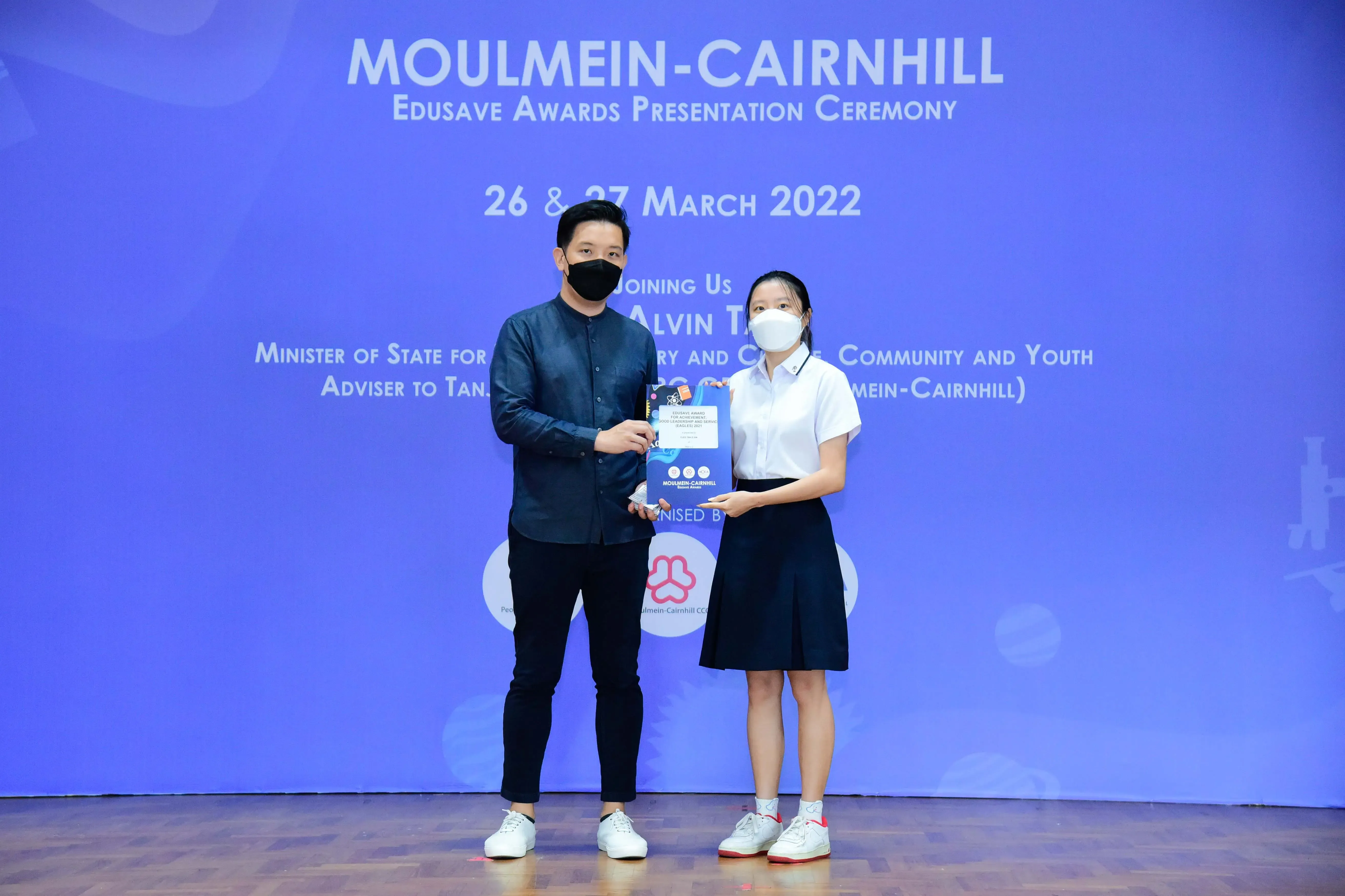 Two masked people hold a certificate on stage at the Moulmein-Cairnhill Edusave Awards Ceremony, March 2022.