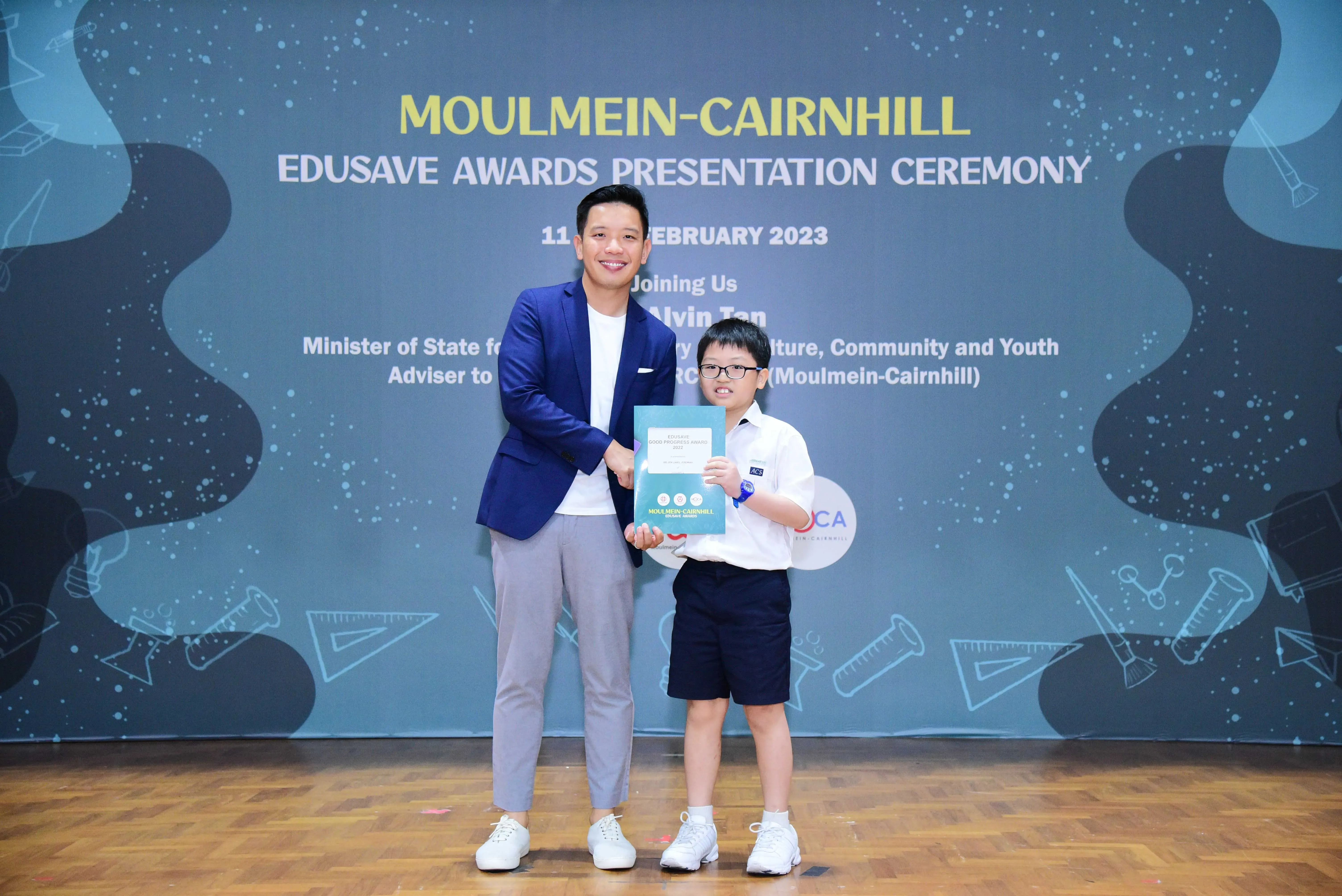 A man in a blue blazer and a young student hold a certificate at the Moulmein-Cairnhill Edusave Awards Ceremony 2023.