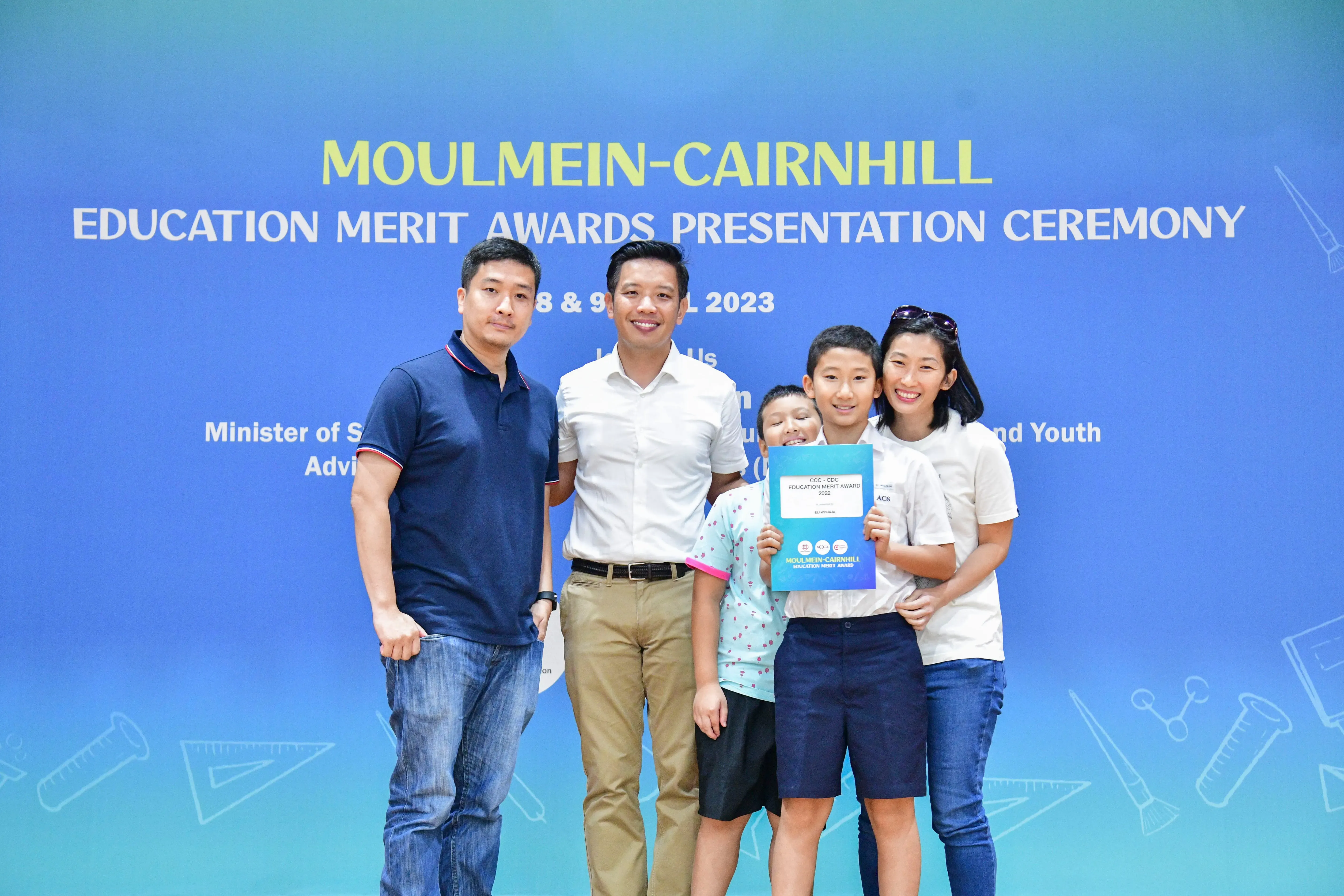 Five people pose before a Moulmein-Cairnhill Education Merit Awards 2023 banner, with a boy holding a certificate.