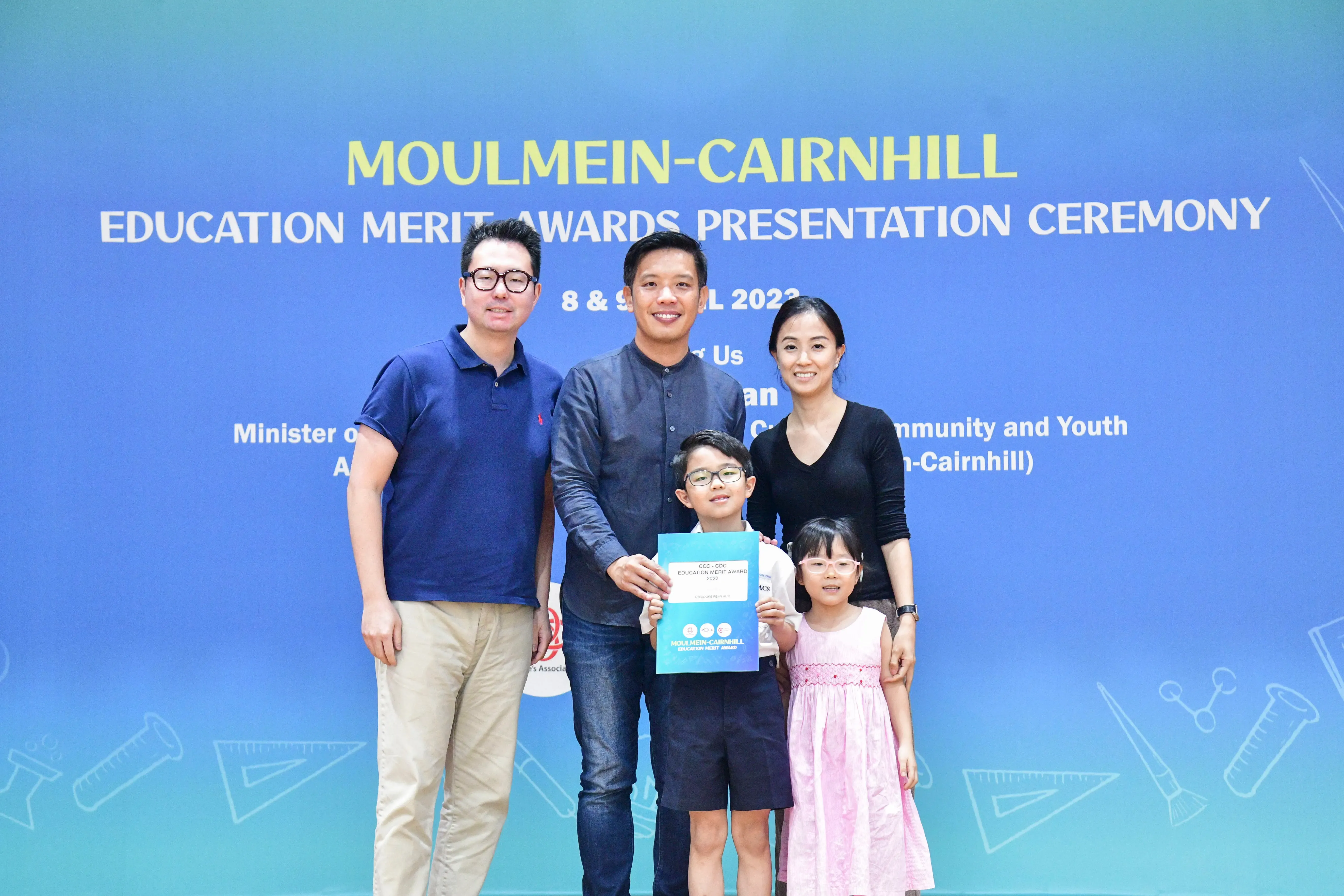 Five people pose before a Moulmein-Cairnhill Education Merit Awards banner; a boy holds a certificate.