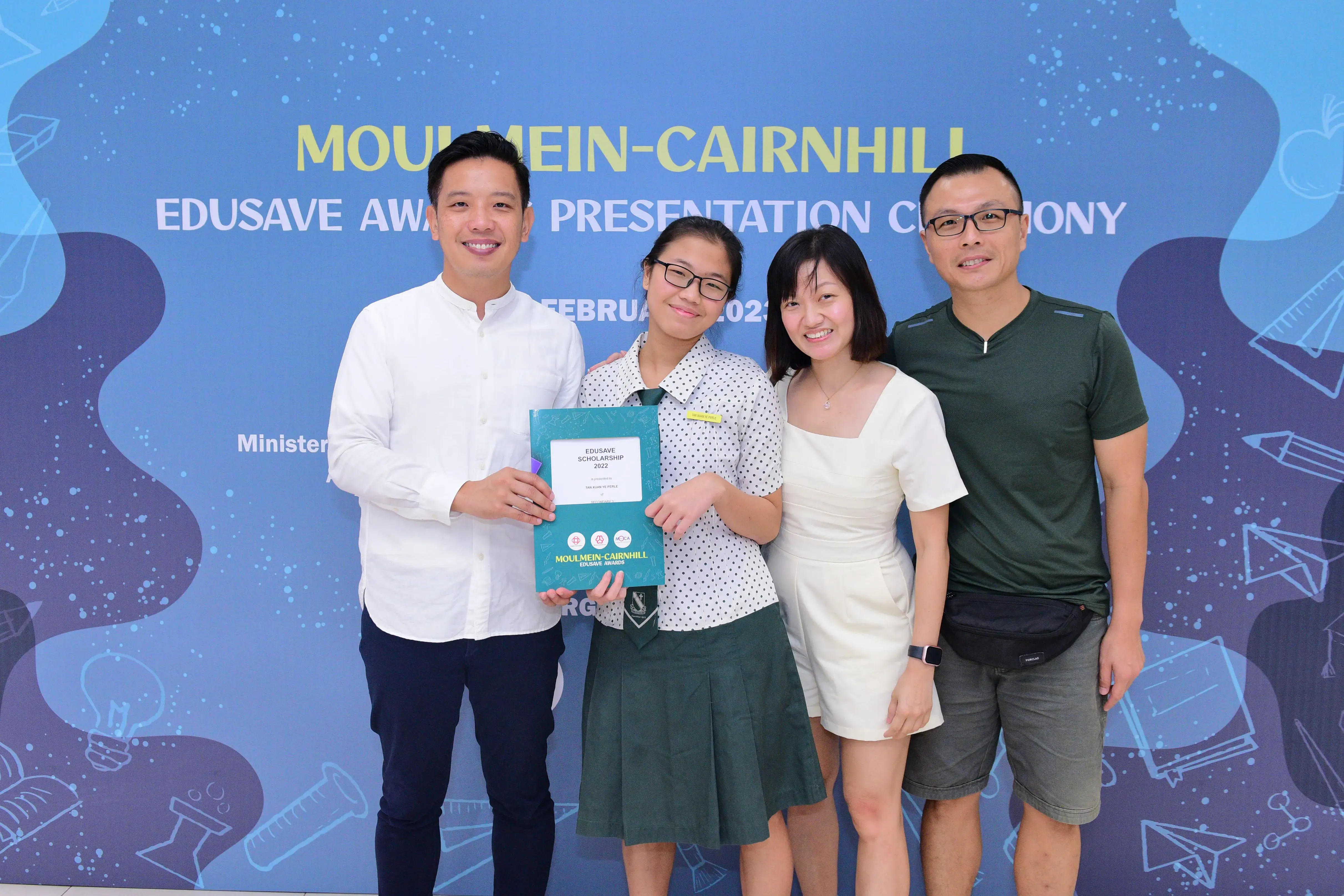 Four people smile at the Moulmein-Cairnhill Edusave Awards Ceremony, with a student holding a 2022 scholarship certificate.