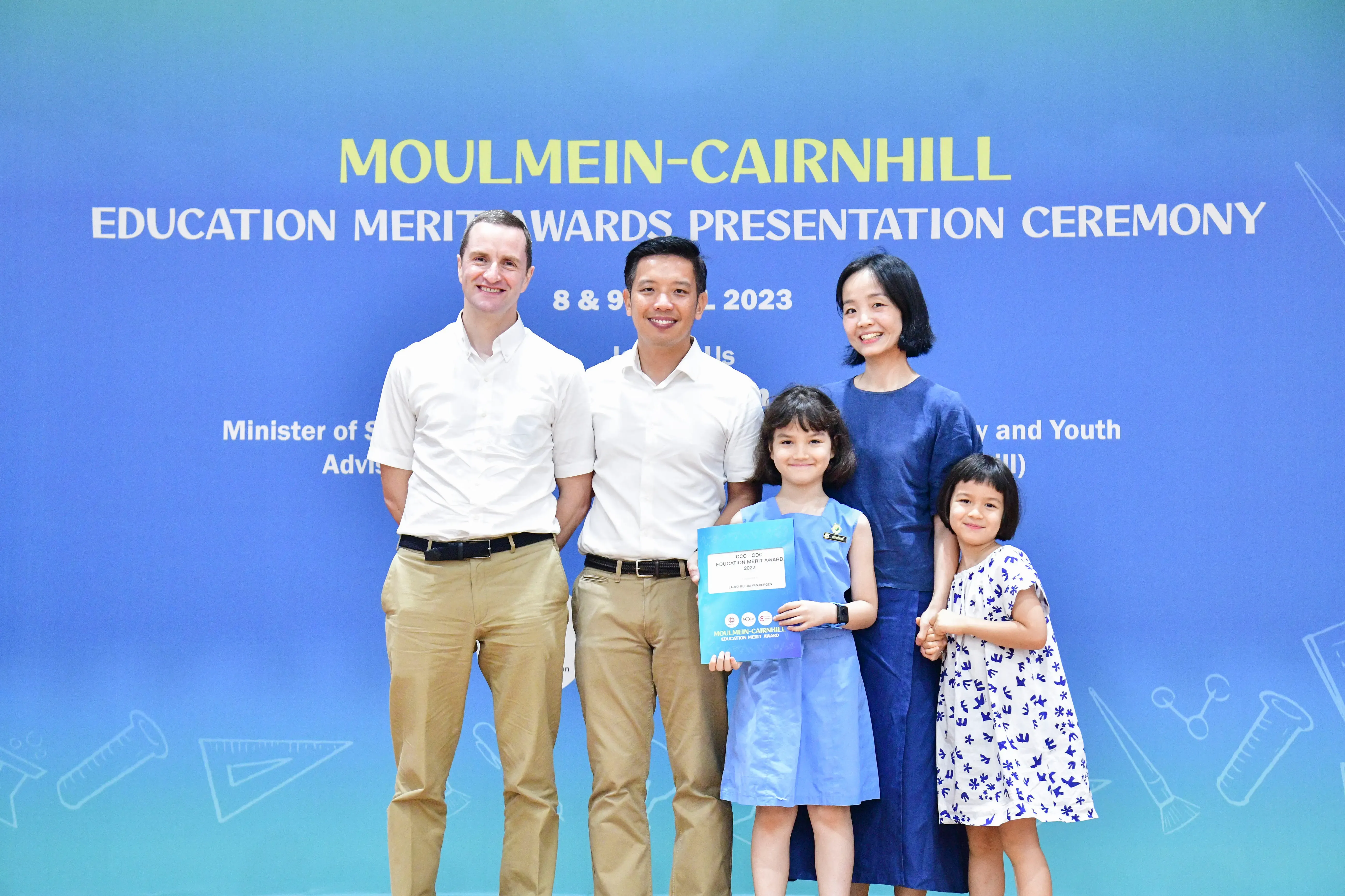 Five people pose in front of a blue Moulmein-Cairnhill Education Merit Awards 2023 banner, a girl holding an award.