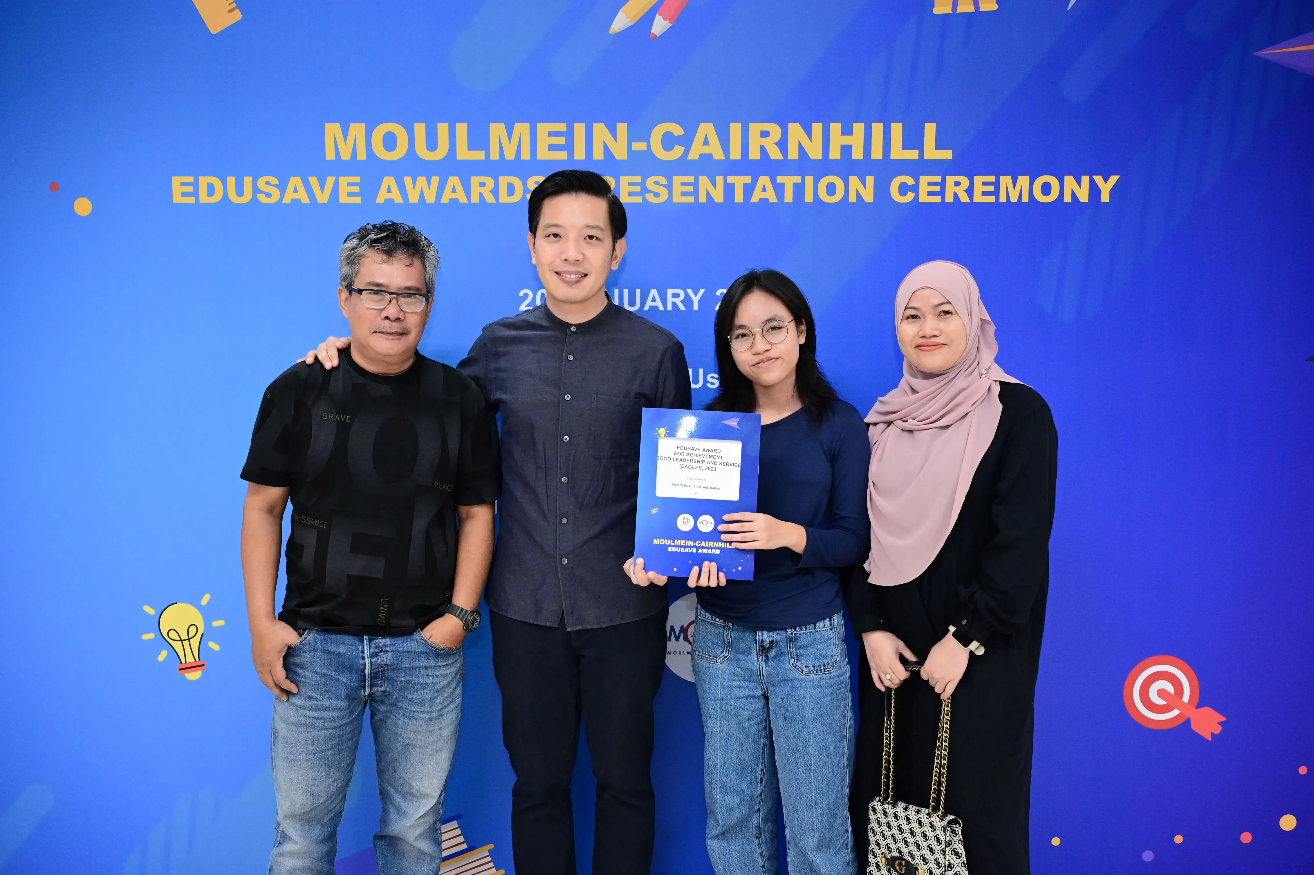 Four people pose in front of a Moulmein-Cairnhill Edusave Awards Presentation Ceremony banner, one holding a certificate.