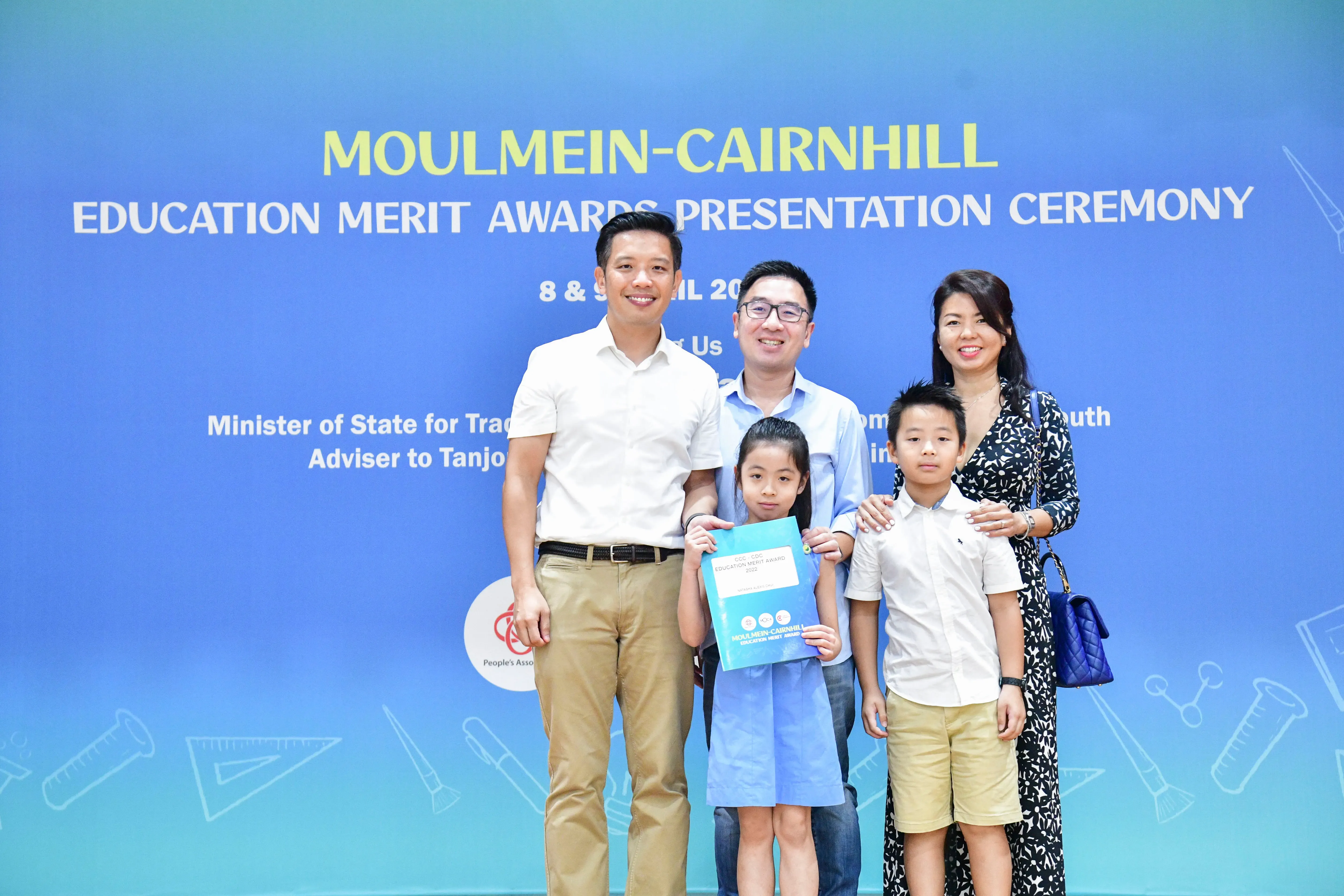 Three adults and two children pose smiling before a Moulmein-Cairnhill Education Merit Awards Ceremony banner.