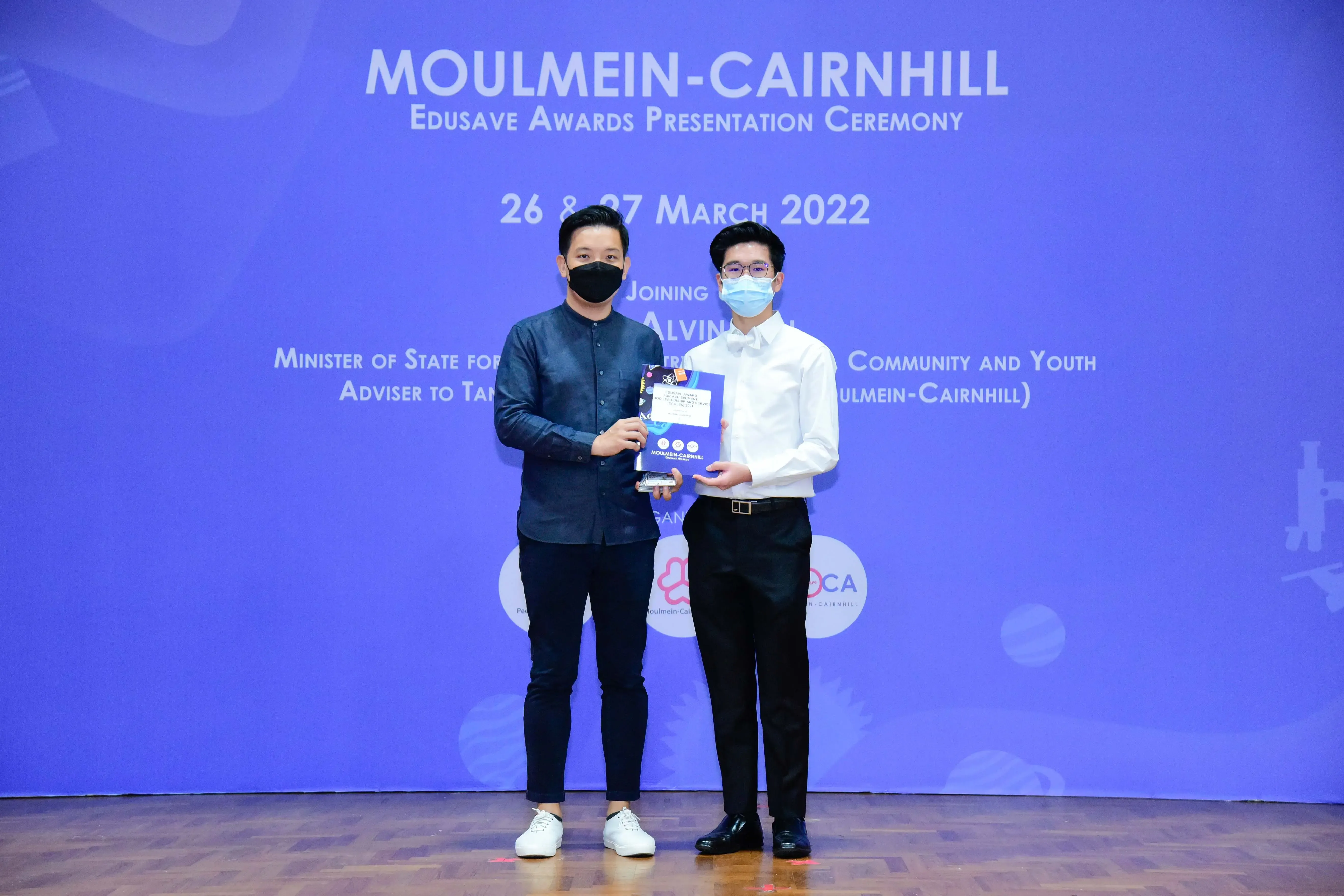 Two masked men hold an award at the Moulmein-Cairnhill Edusave Awards Presentation Ceremony, March 2022.