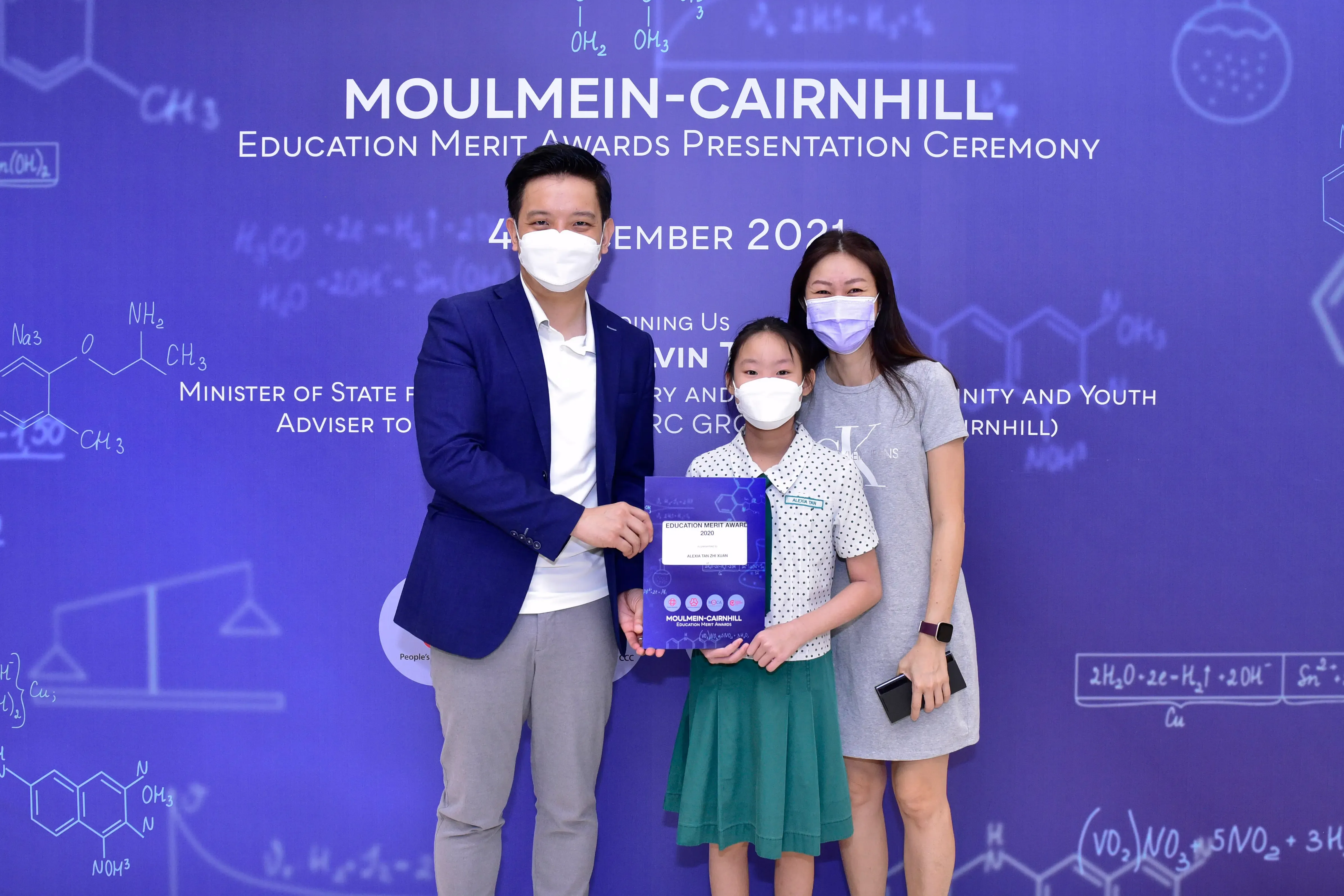 Three masked people pose at the Moulmein-Cairnhill Education Merit Awards Ceremony 2021, with a girl holding an award certificate.