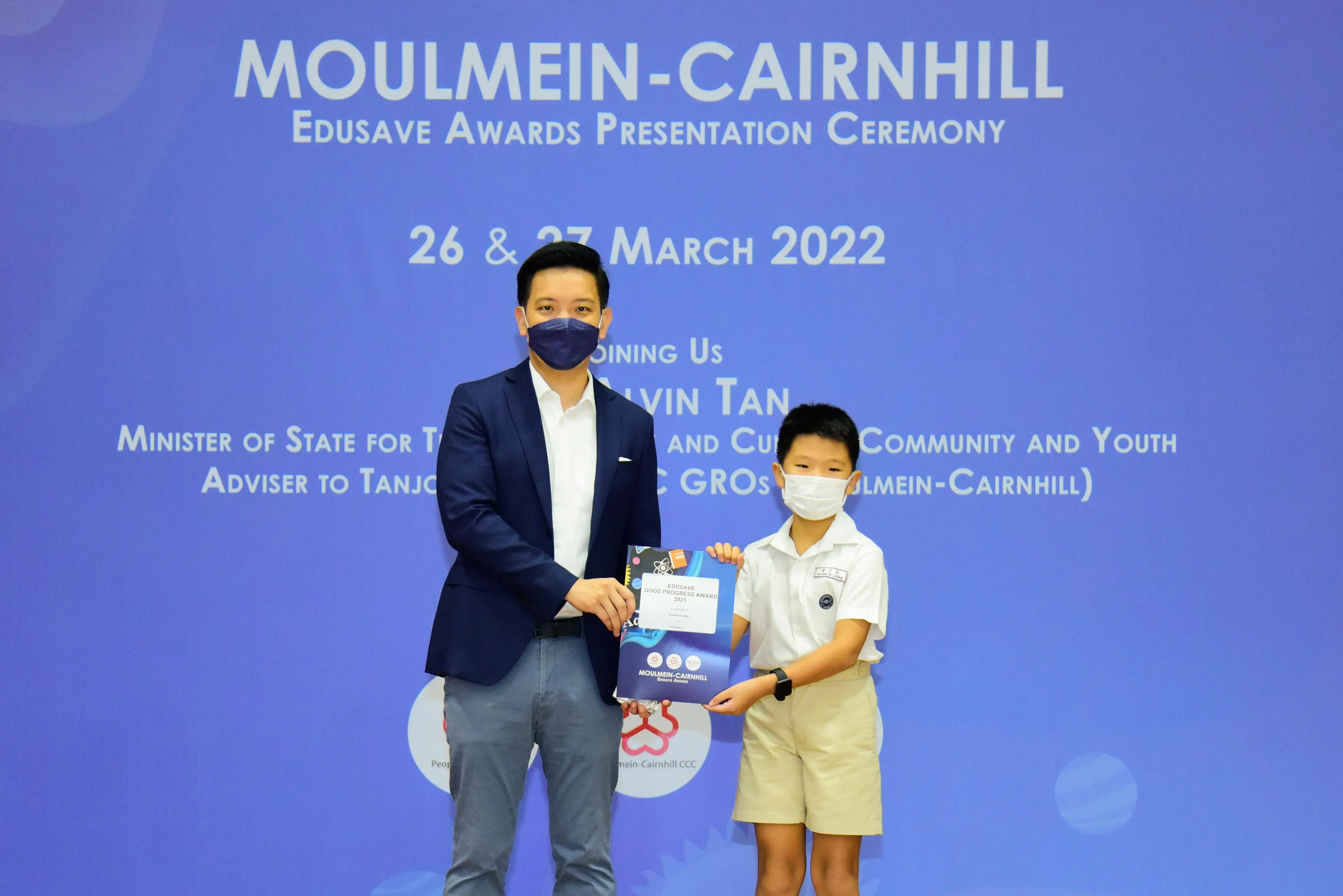 An adult and a young student in masks hold an award certificate at the Moulmein-Cairnhill Edusave Awards Ceremony 2022.