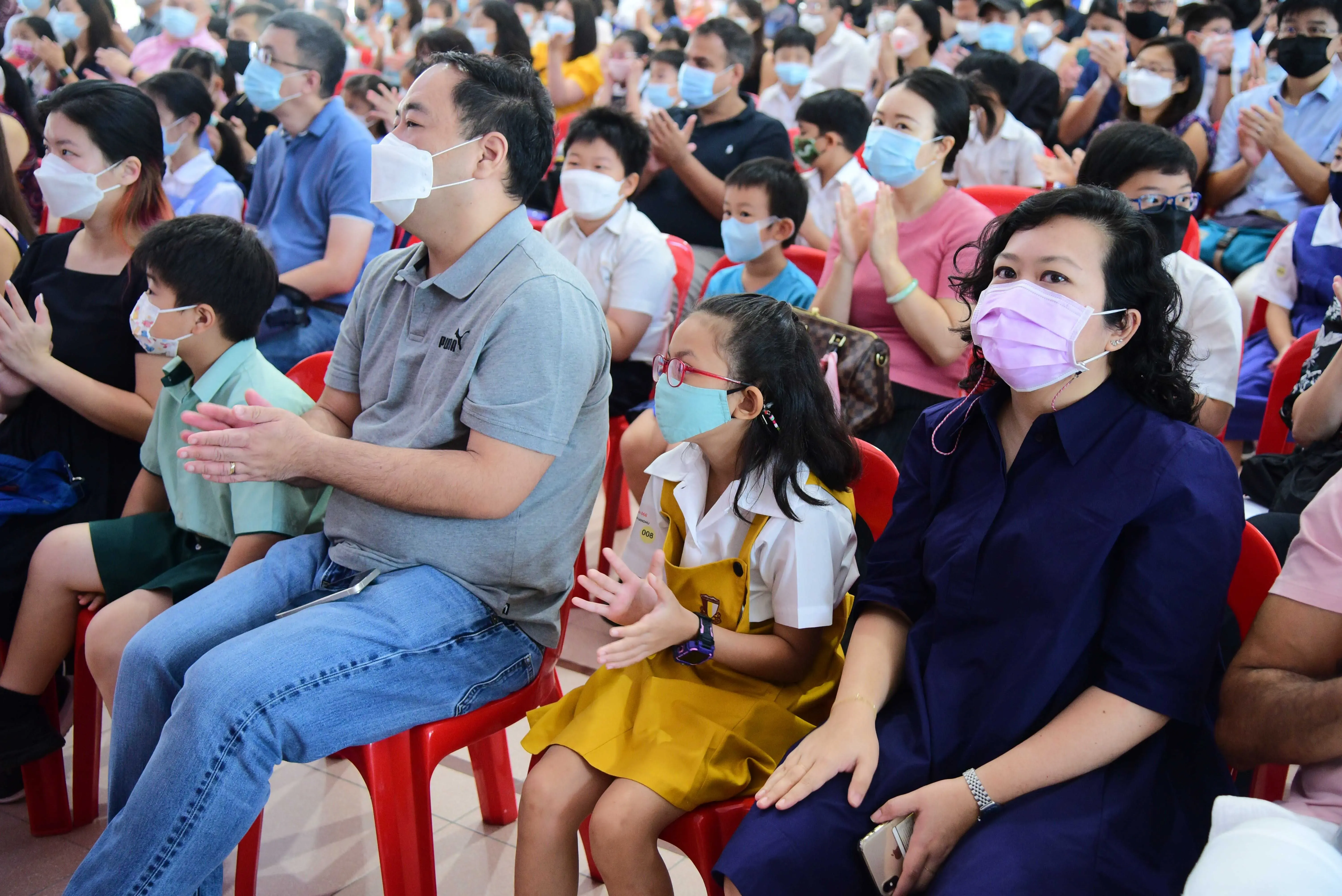 A large crowd of adults and children wearing face masks sit on red chairs at an indoor event, many clapping.