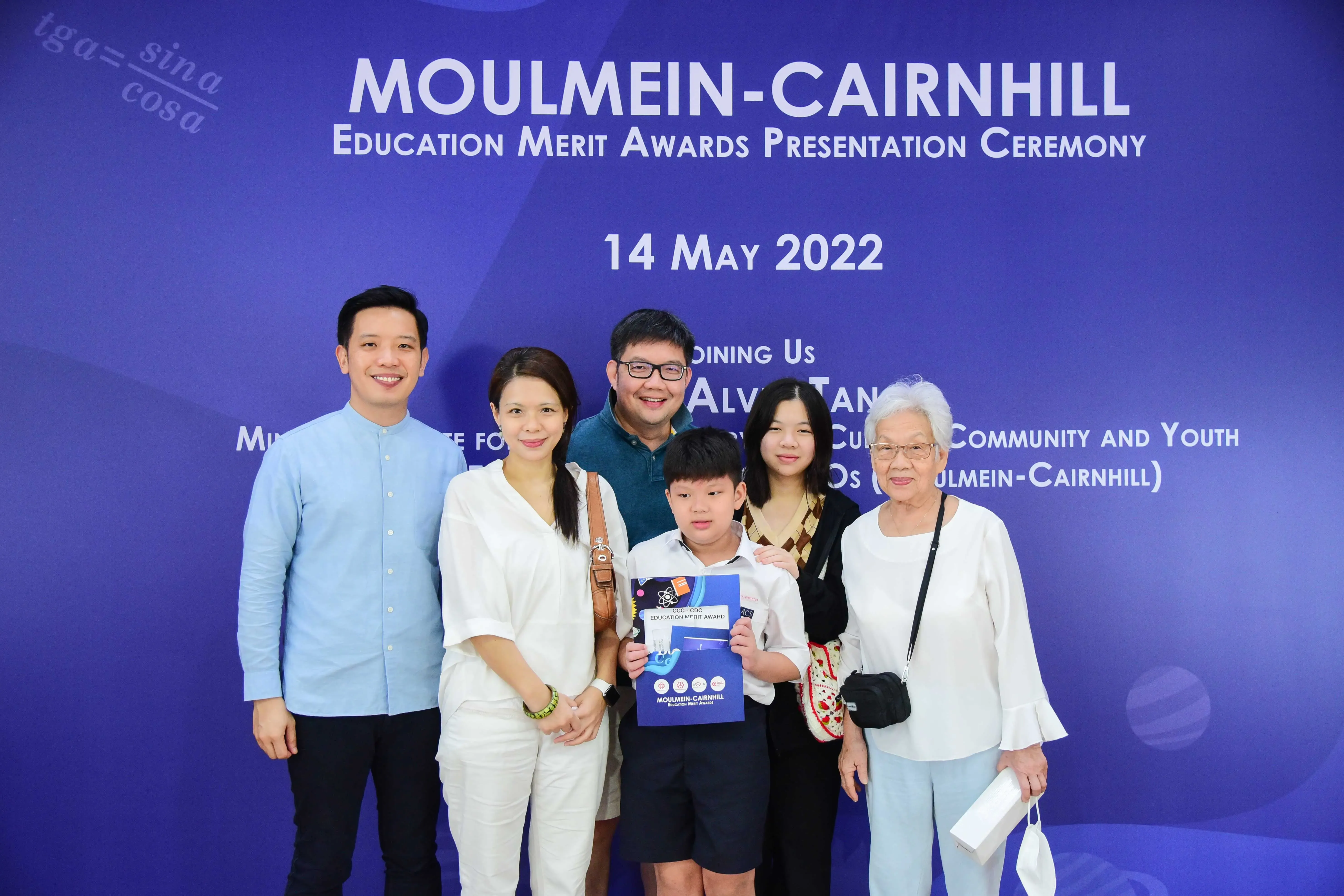 Five adults and a young boy pose before a Moulmein-Cairnhill Education Merit Awards banner dated 14 May 2022.