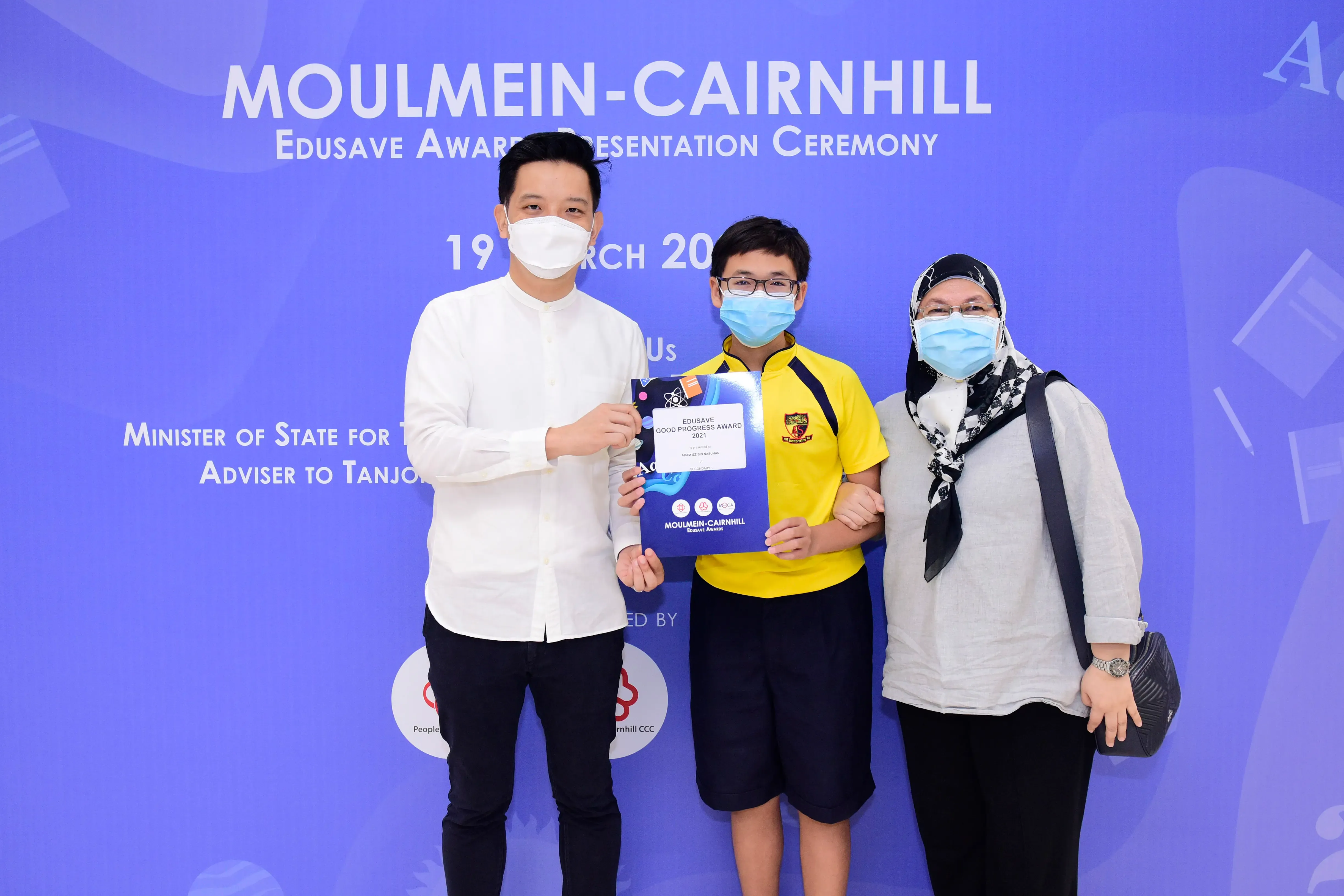 Three masked people pose at the Moulmein-Cairnhill Edusave Award Presentation Ceremony, with a boy holding an award folder.