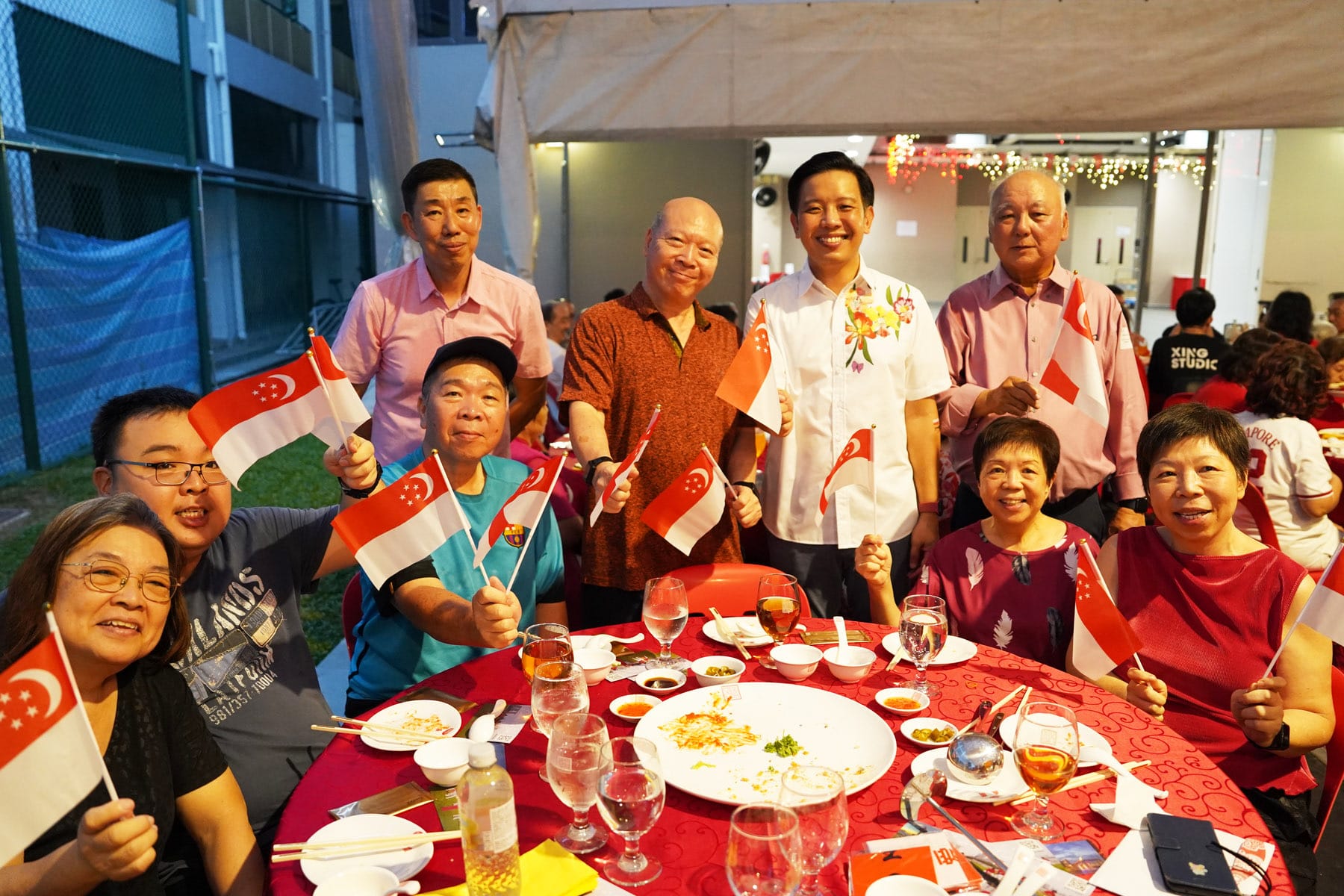 A group of smiling people hold Singapore flags around a red dinner table with food and glasses at an outdoor event.