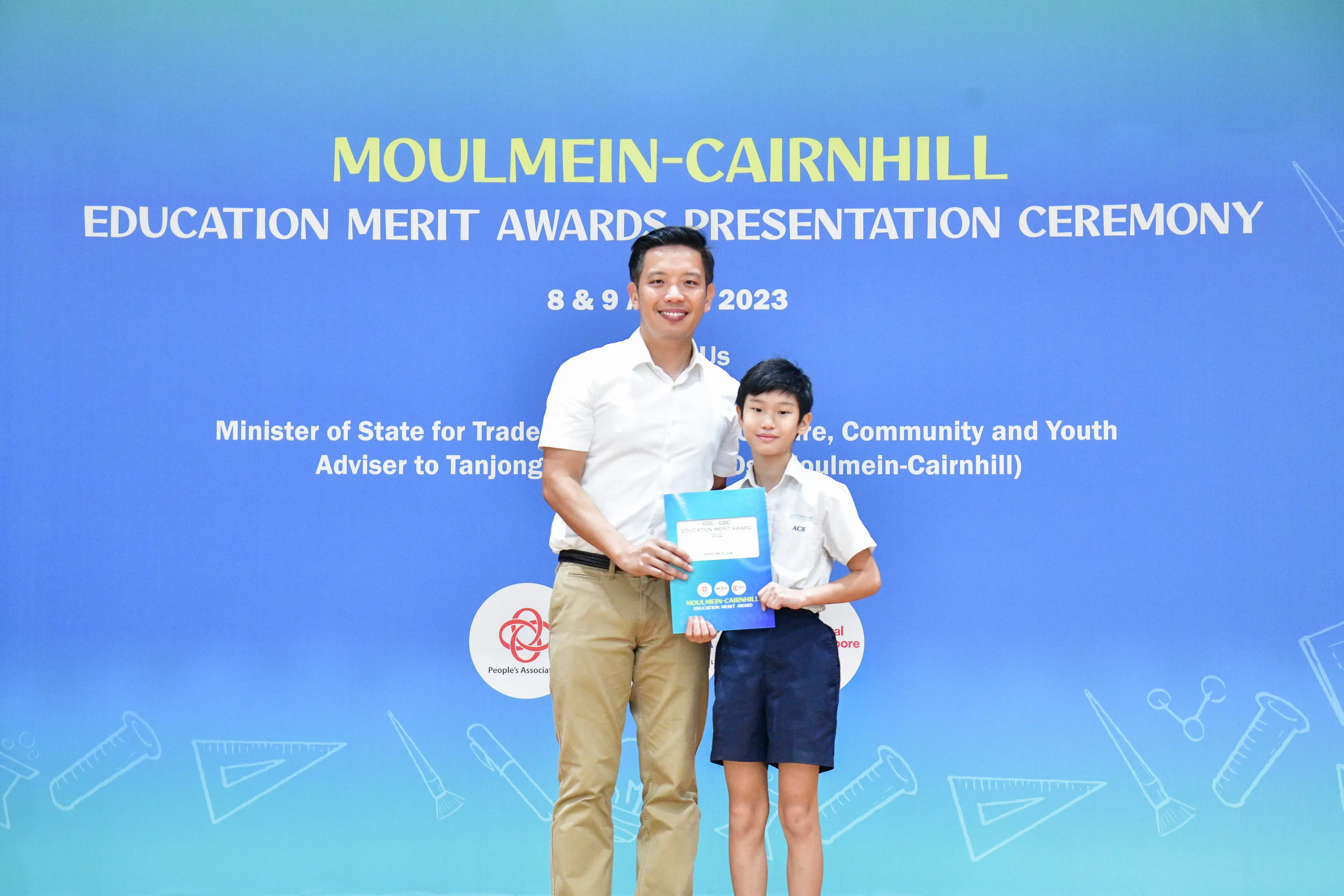 An adult man and a young boy hold an award together in front of a Moulmein-Cairnhill Education Merit Awards banner.