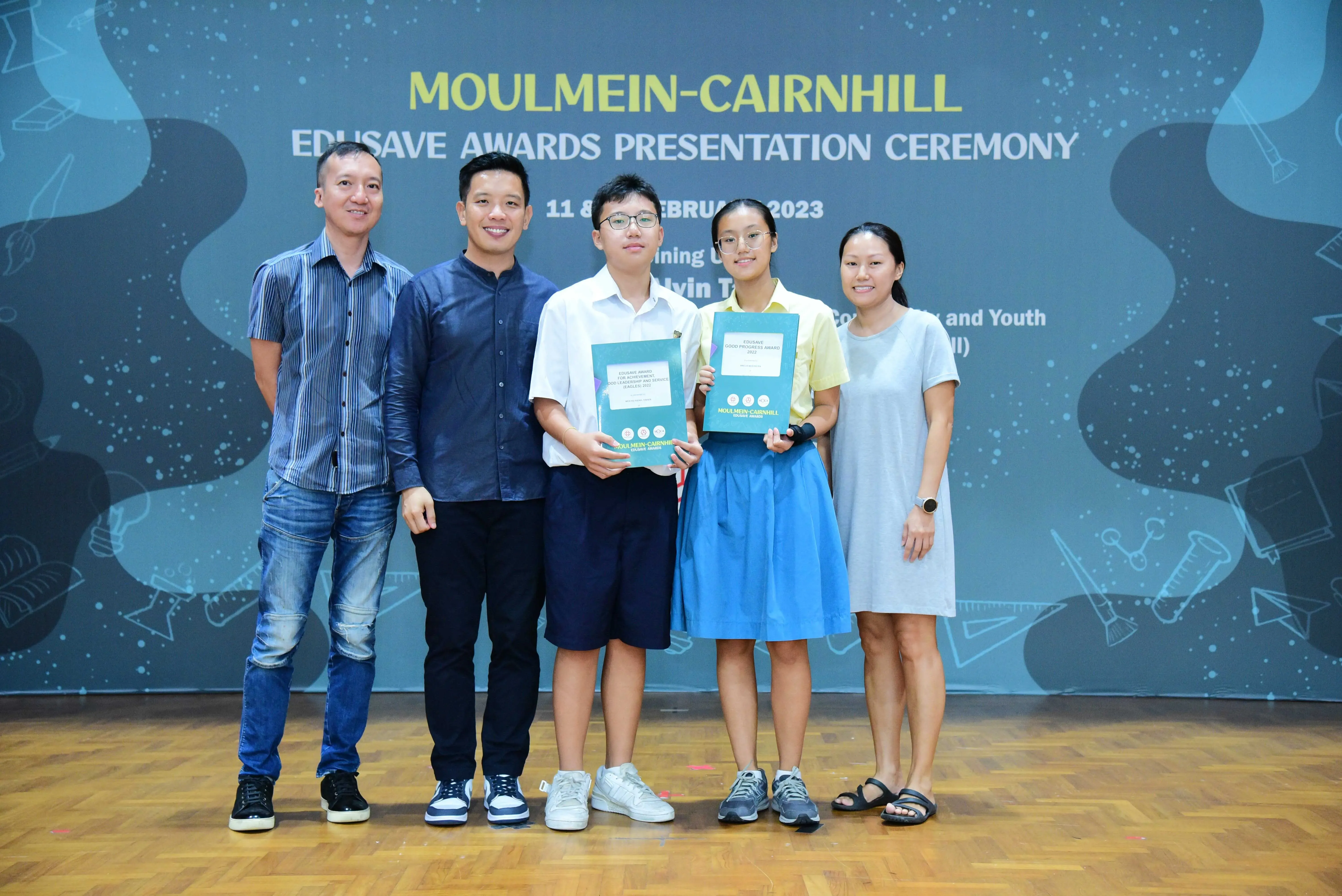 Five people pose before a Moulmein-Cairnhill Edusave Awards Presentation Ceremony banner; two students hold award folders.