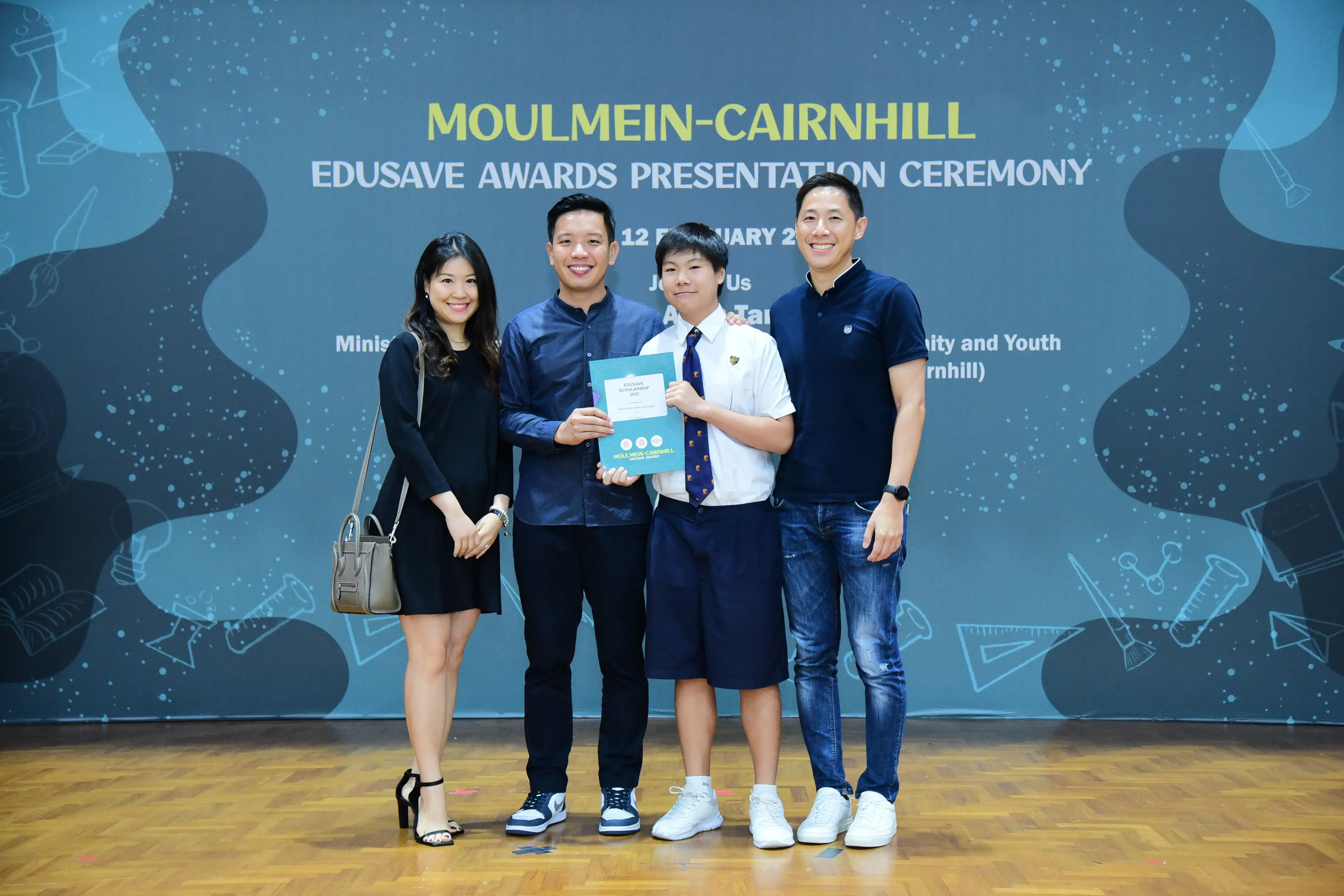 Four people pose smiling at the Moulmein-Cairnhill Edusave Awards Presentation Ceremony, with a student holding a certificate.