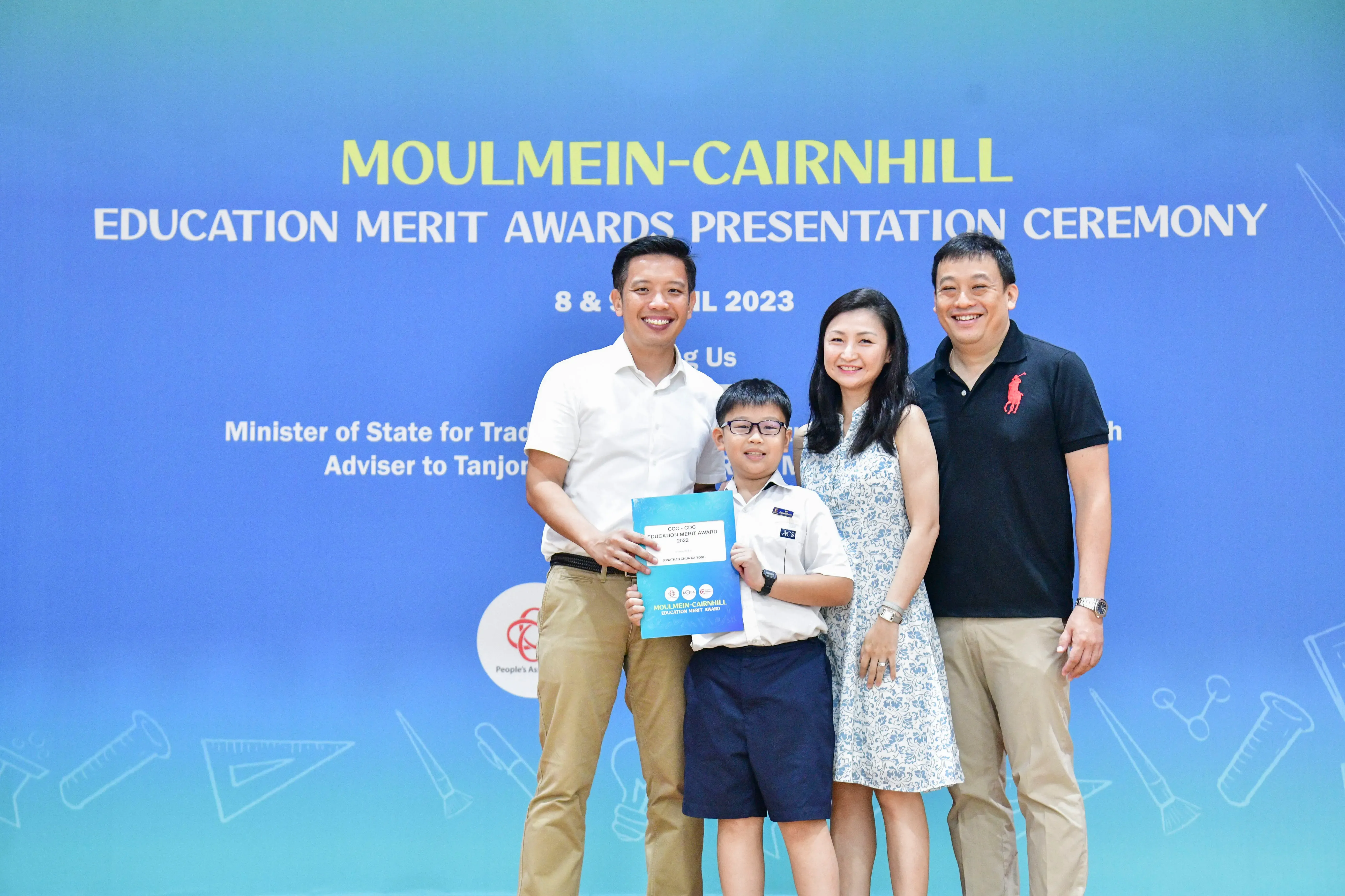 Three adults and a boy holding an award folder pose in front of a Moulmein-Cairnhill Education Merit Awards banner.
