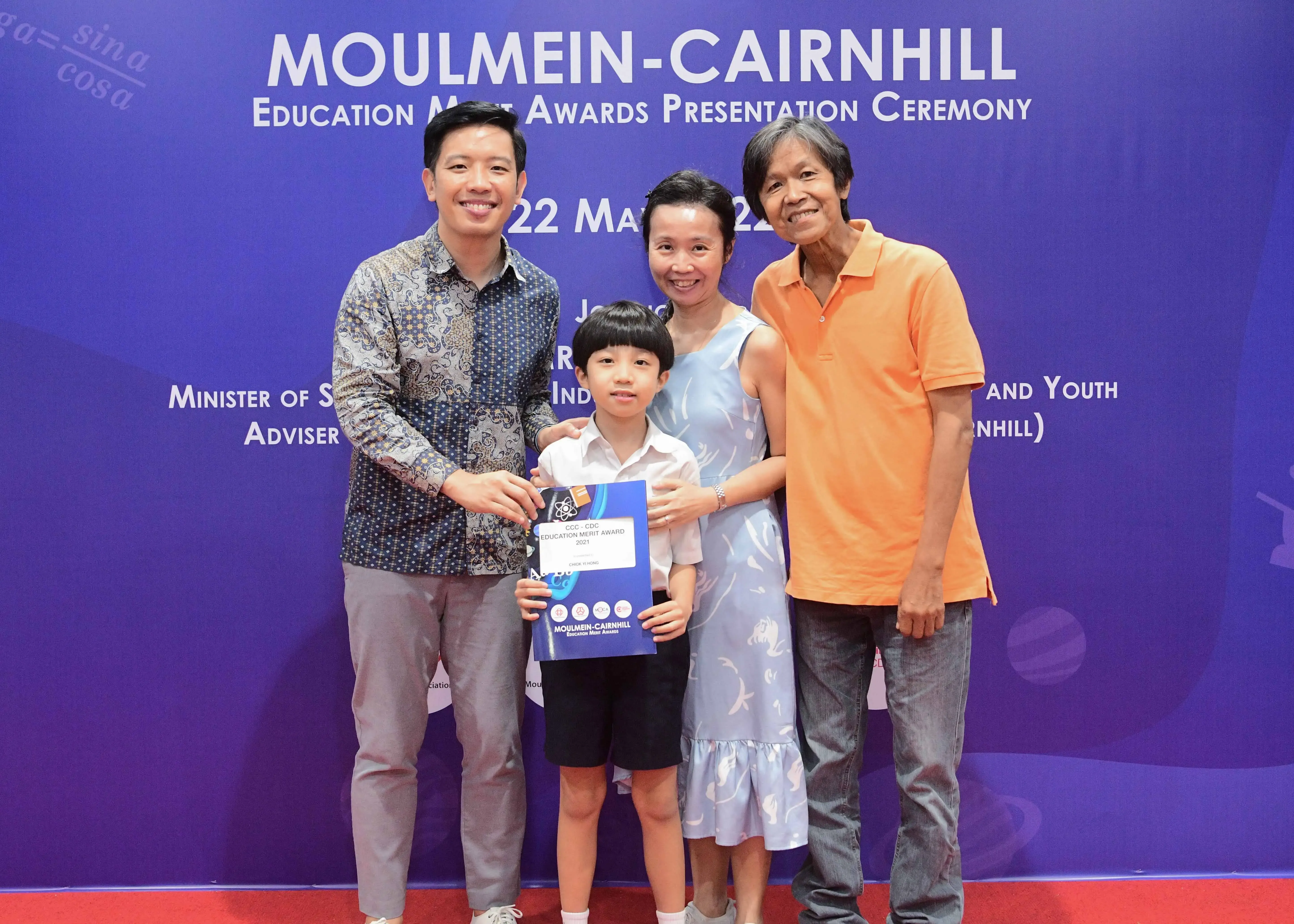 Four people pose before a Moulmein-Cairnhill Education Merit Awards banner, with a boy holding a blue award certificate.