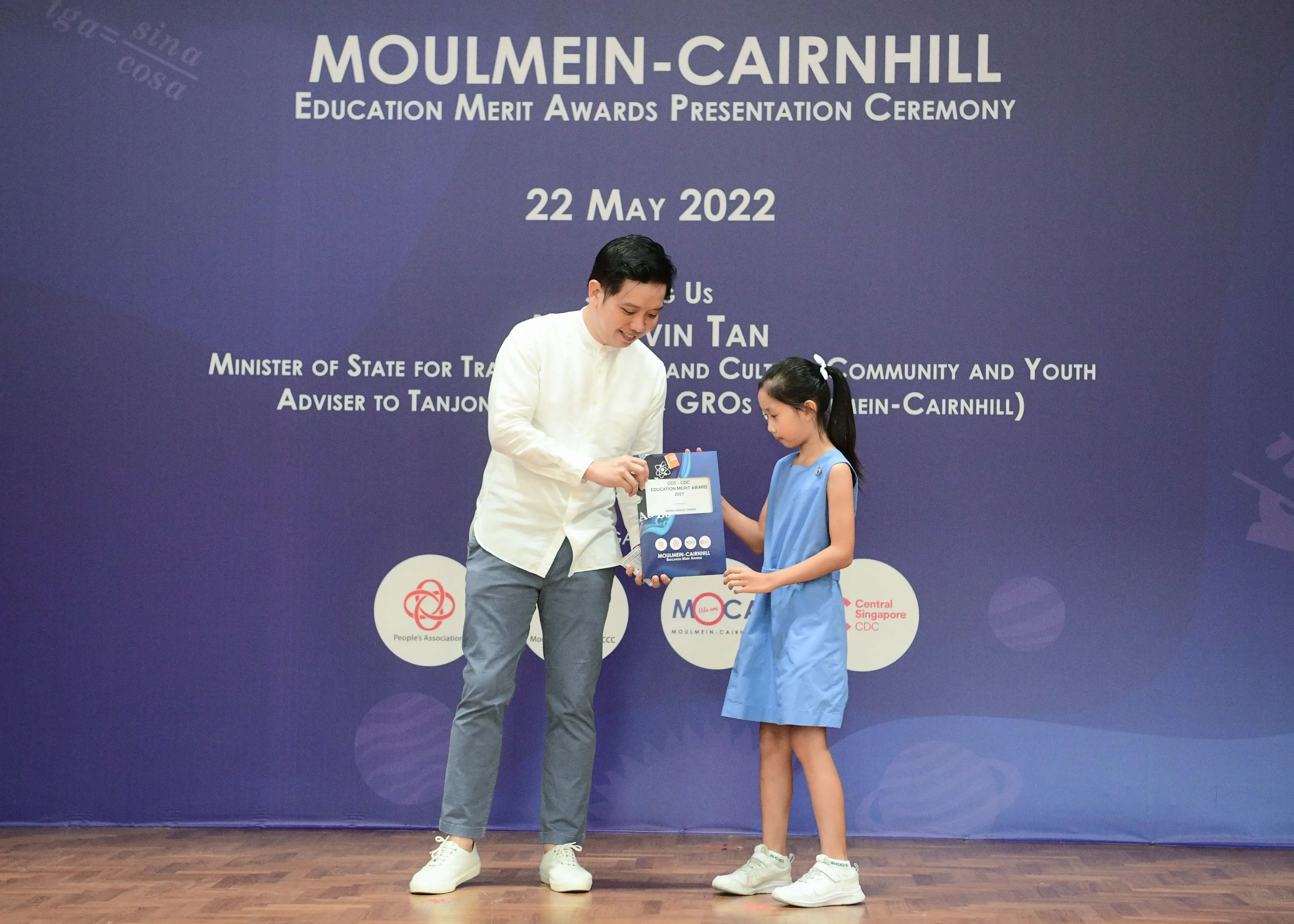 A man in white hands an award packet to a young girl in a blue dress at the Moulmein-Cairnhill Education Merit Awards, May 2022.