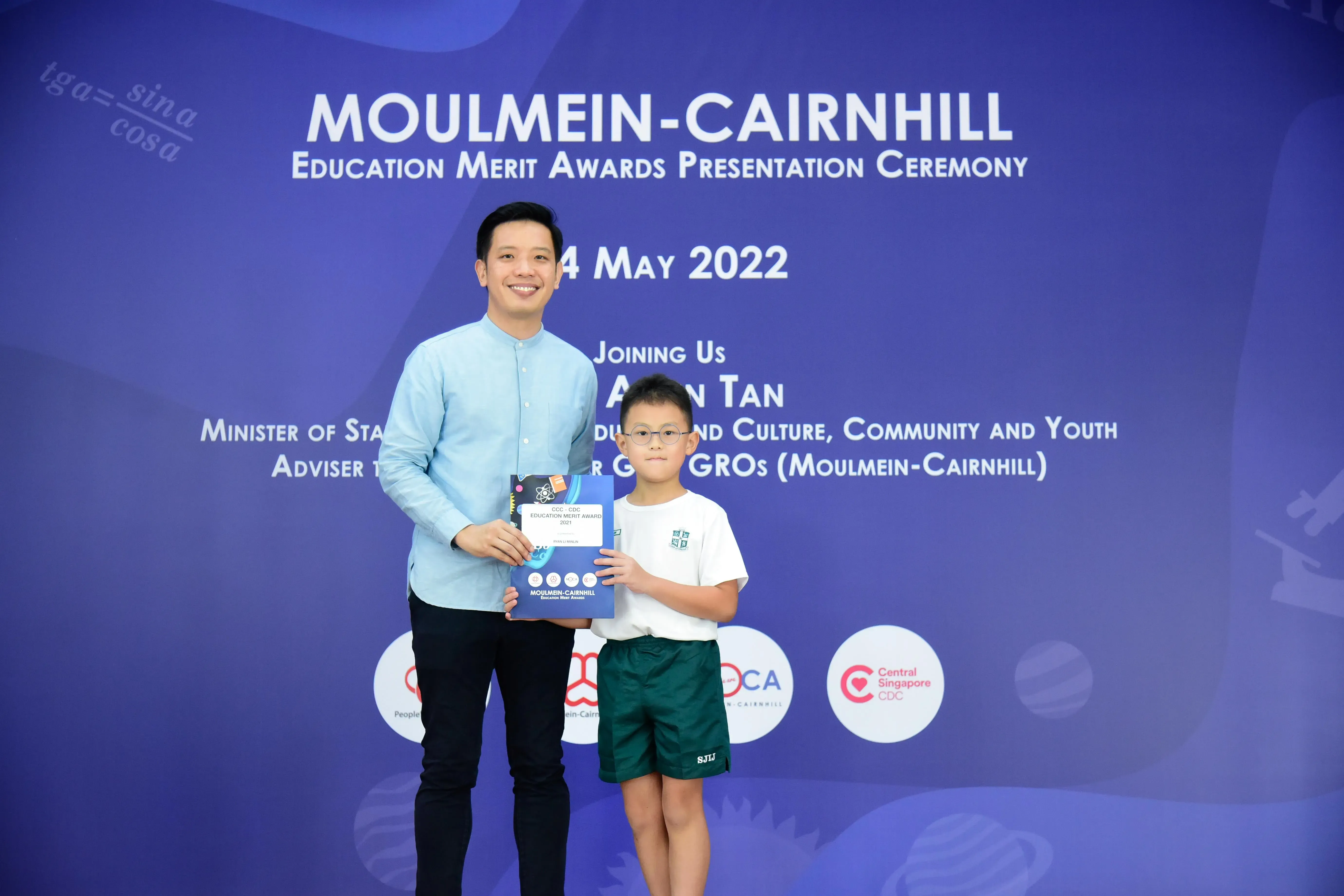 A man and a young boy holding award booklets stand before a Moulmein-Cairnhill Education Merit Awards banner dated 4 May 2022.
