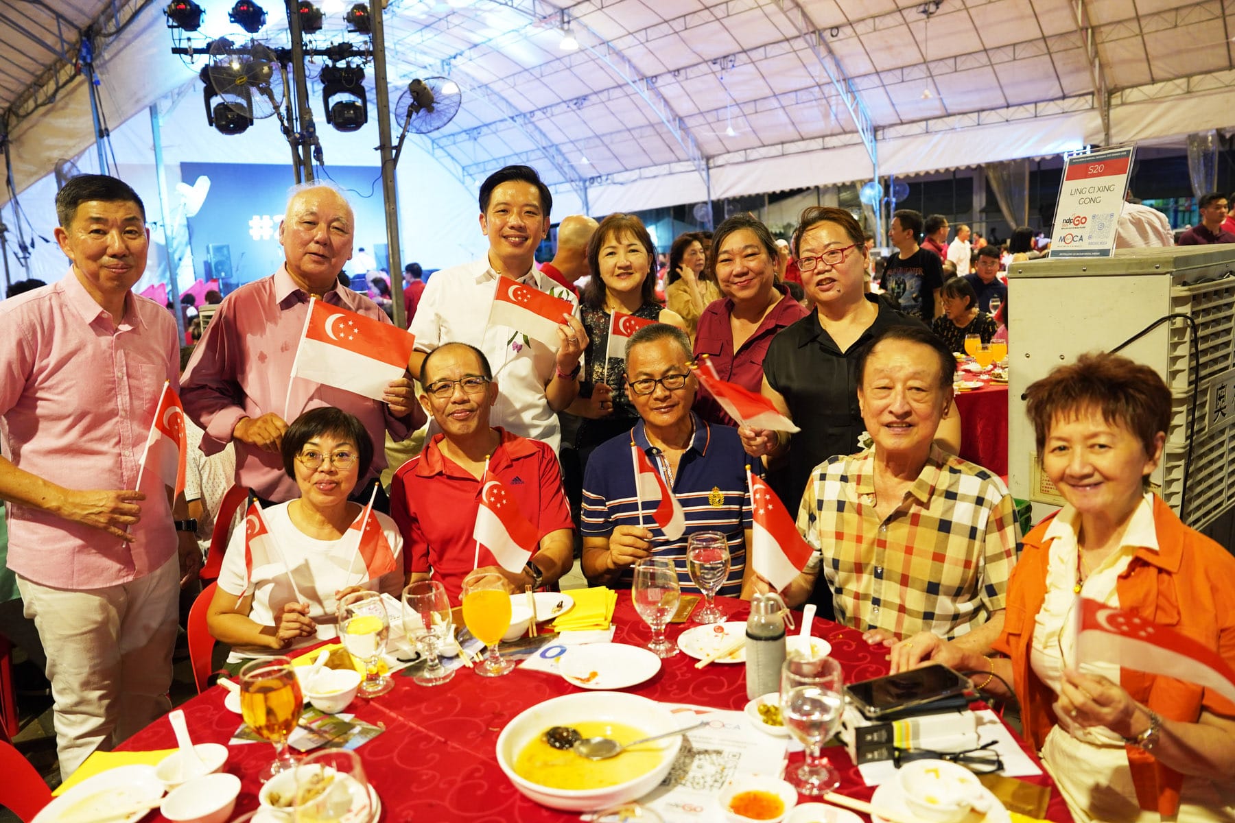 Group of people smiling and holding small Singapore flags at a festive dinner under a tent.