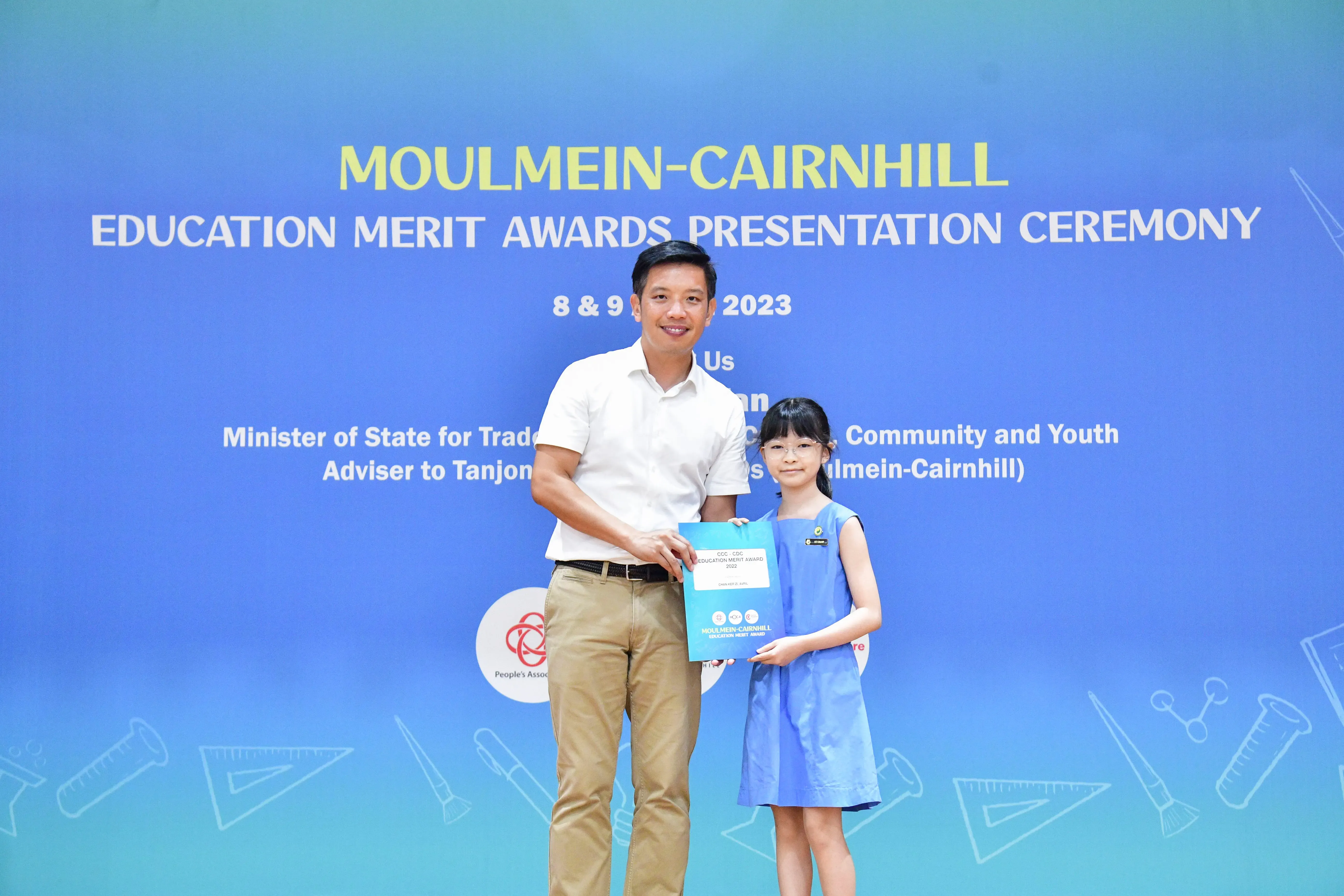 A man and a young girl hold a certificate together in front of a Moulmein-Cairnhill Education Merit Awards banner.